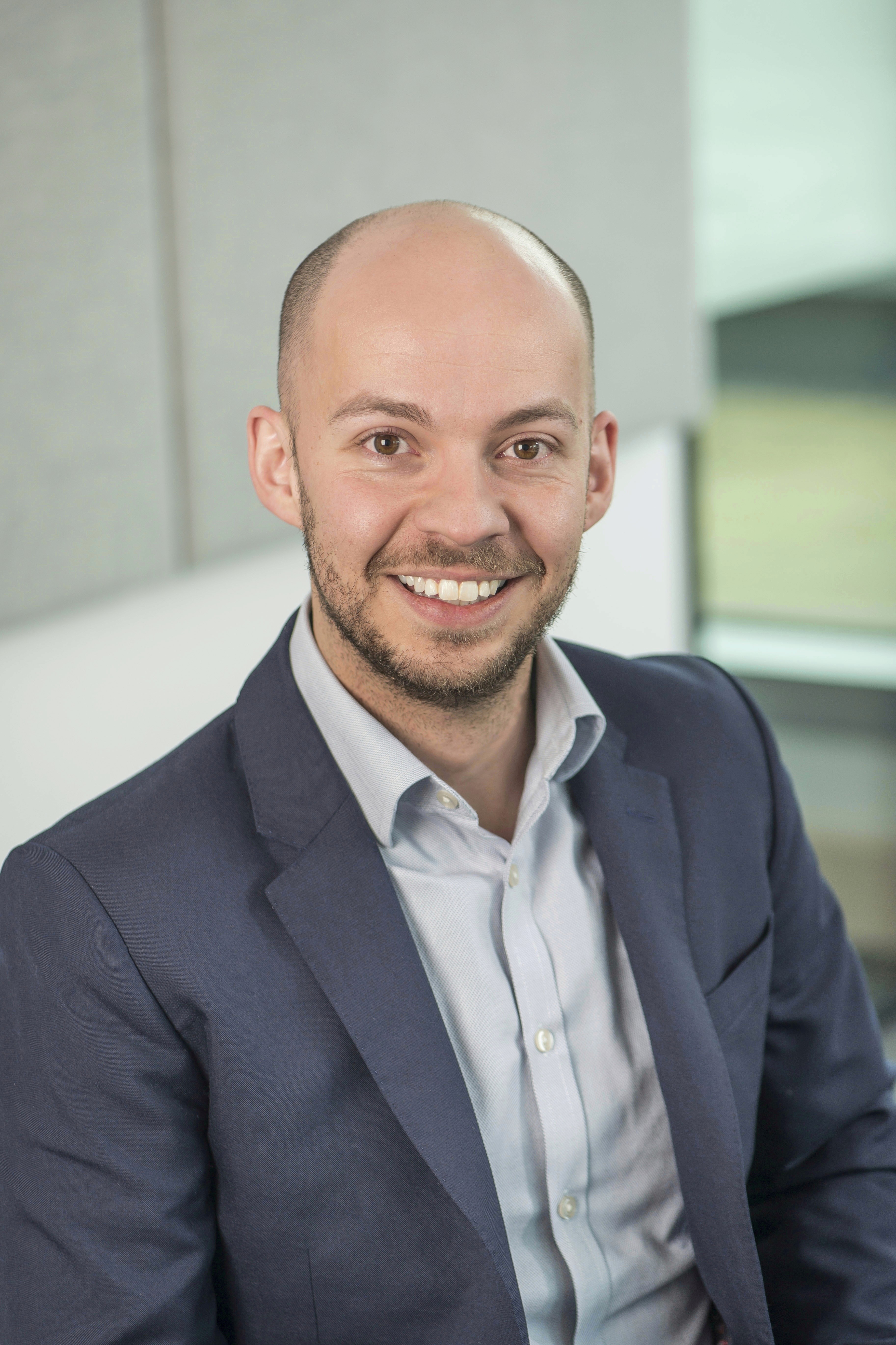 Mature man with glasses and a trimmed beard smiling confidently, standing in warm natural light against a modern backdrop.