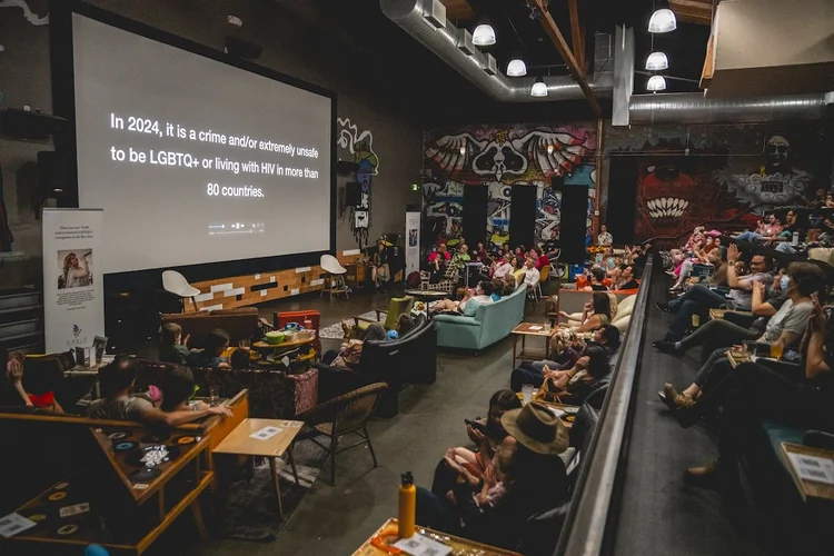 Em um espaço interno aconchegante e eclético, repleto de murais coloridos, um grupo de pessoas observa atentamente uma grande tela de projeção exibindo uma mensagem sobre os direitos LGBTQ+ e a criminalização do HIV em 2024.