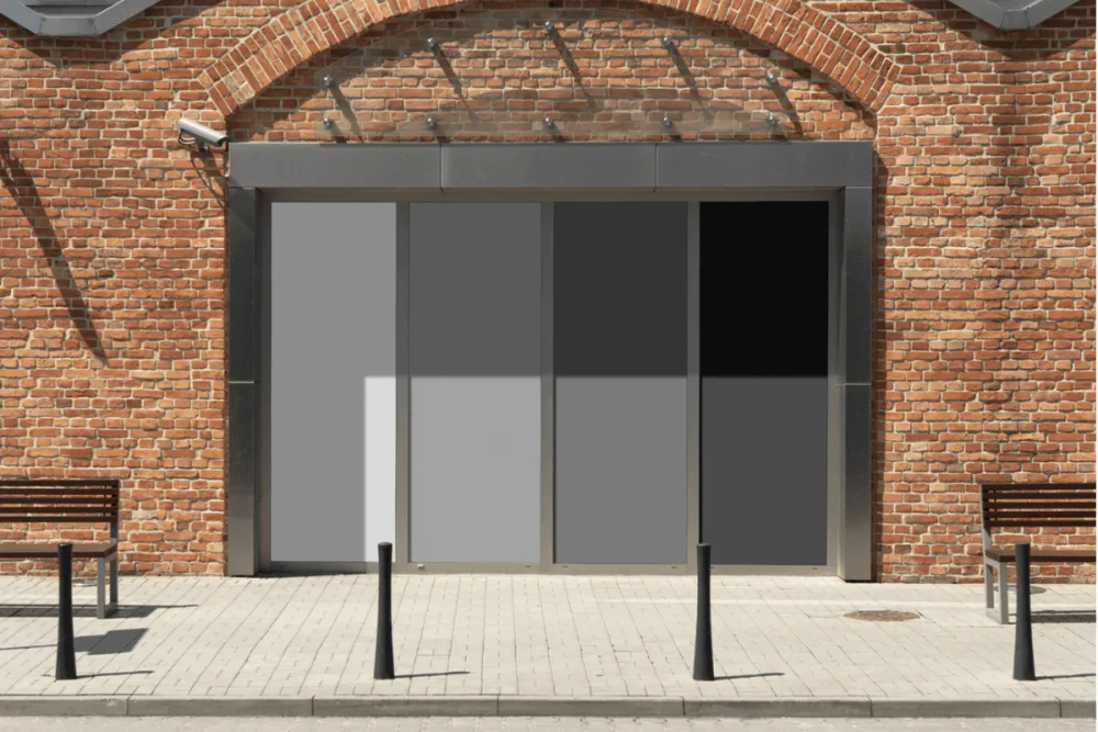 Facade mockup featuring a modern glass storefront with large divided panels in grayscale tones, set against a red brick wall. Outdoor setting includes benches and bollards on a paved walkway.