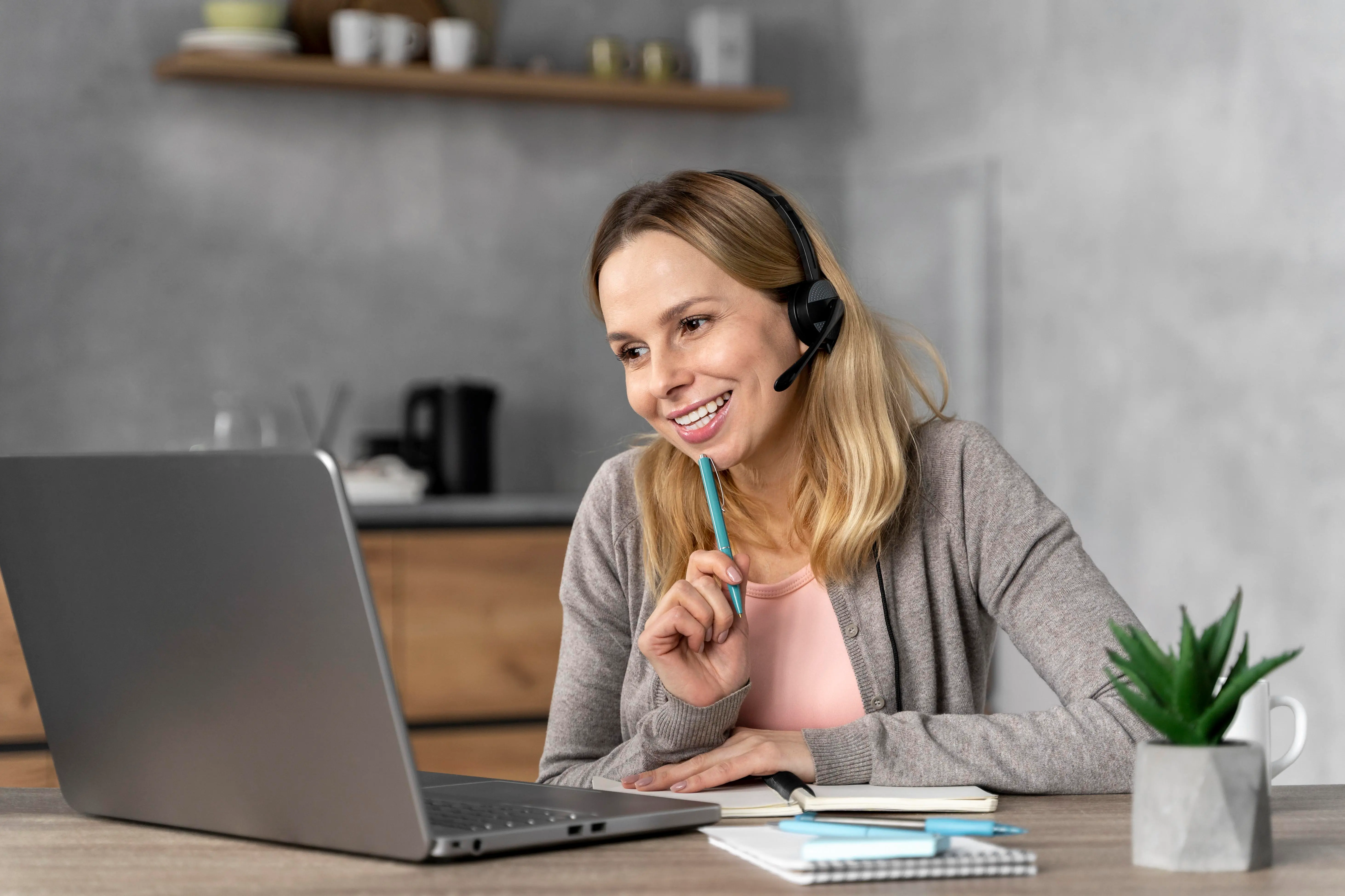 A blonde woman wearing a headset and holding a pen against her chin, with a wide smile looking at a laptop screen