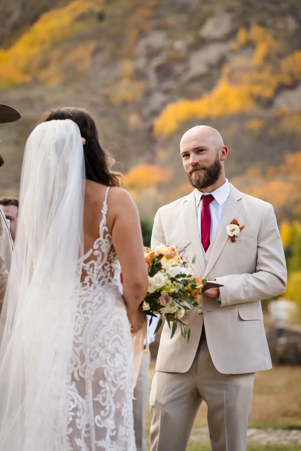 Close-up of a groom in a tan suit looking lovingly at his bride during their wedding vows, with the golden fall colors of the Colorado mountains softly blurred in the background.