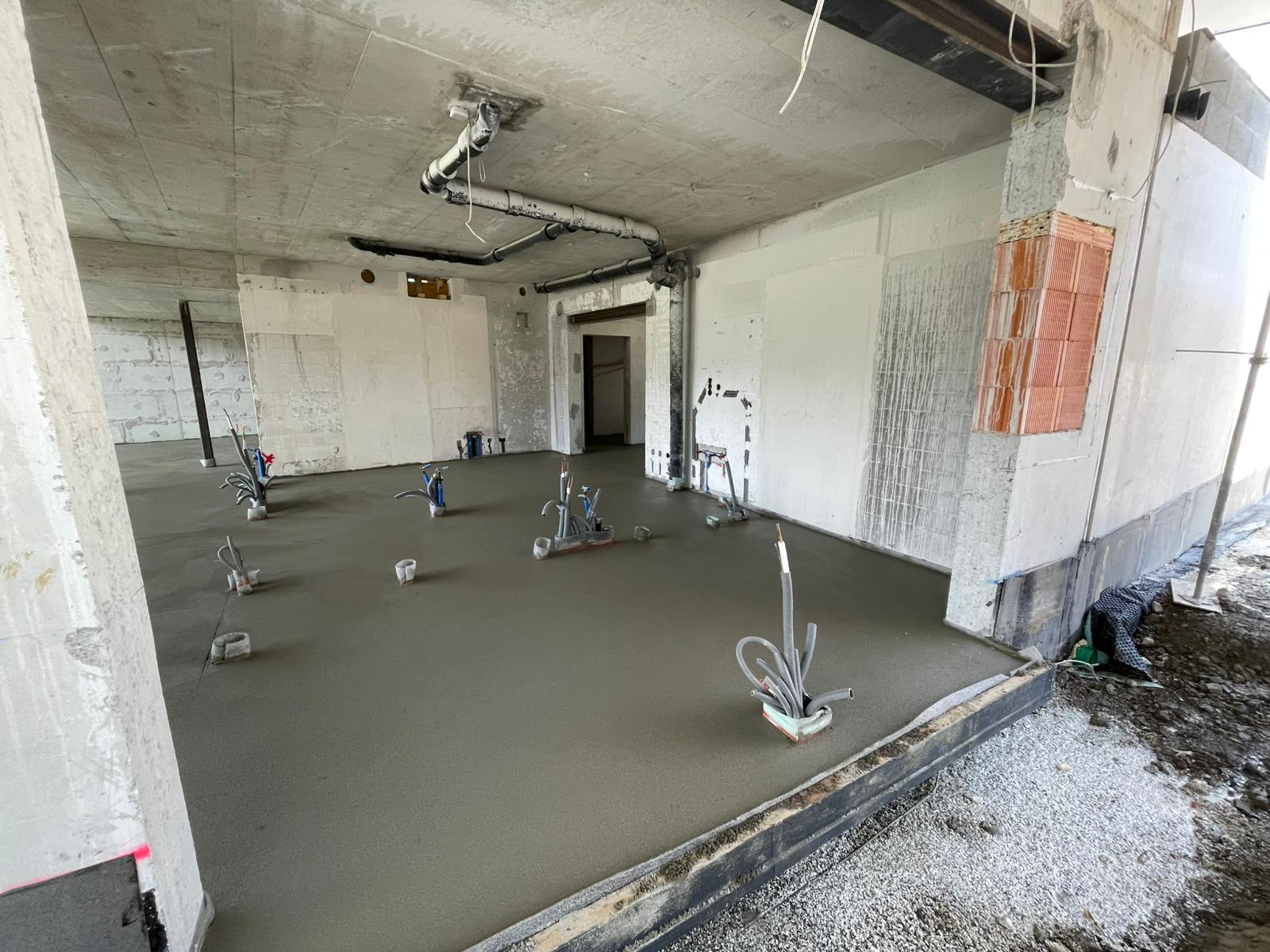 Eine Baustelle mit neuem Betonboden, Rohren und unfertigen Wänden zeigt den Fortschritt im Gebäudeinneren.
