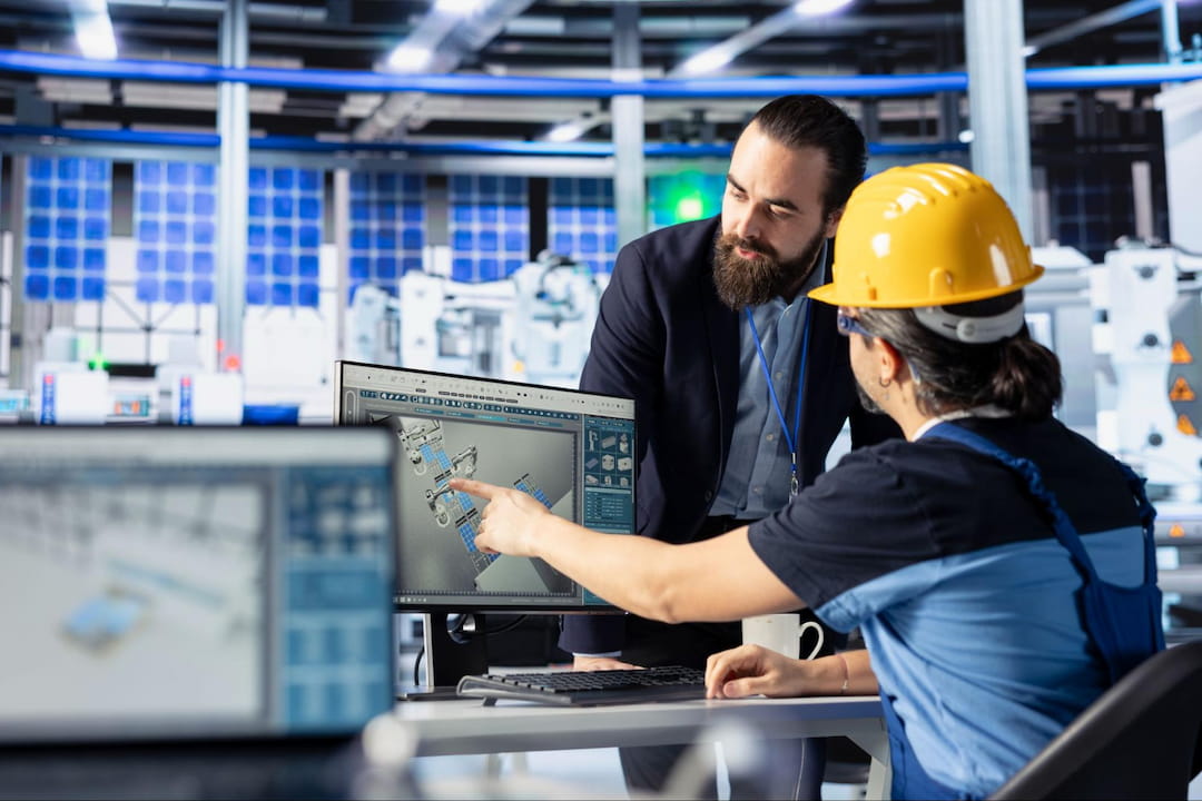 A technician and an engineer in safety gear discuss a CAD design displayed on a computer monitor inside a modern manufacturing facility.