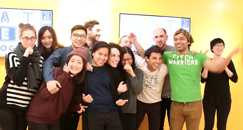 A photo of a group of people standing in an art gallery, in front of a yellow wall with some typographic art work on it.