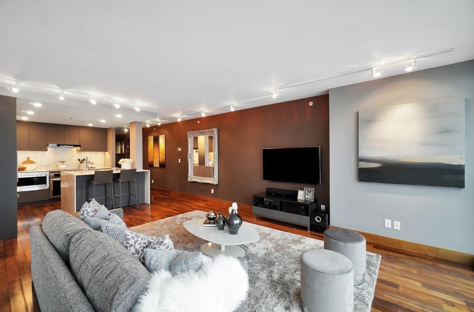 Modern living room featuring a gray sofa, wooden floors, a television, and an open kitchen area in the background.