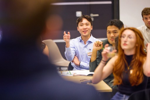 A smiling young man with crossed arms, wearing a plaid shirt and white t-shirt, poses against a dark background.