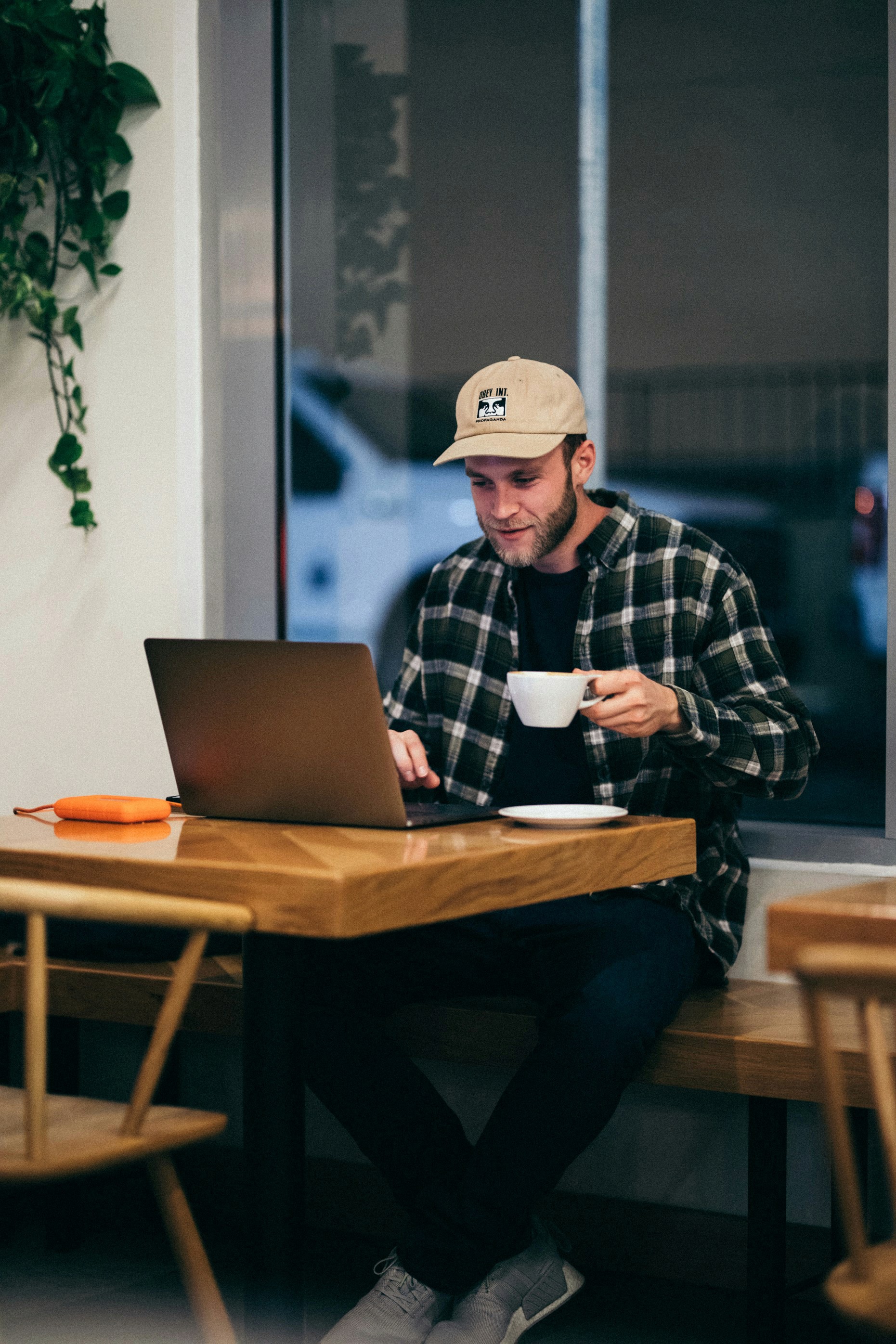 Man wearing a plaid shirt and cap working on a laptop and holding a coffee cup in a cafe.