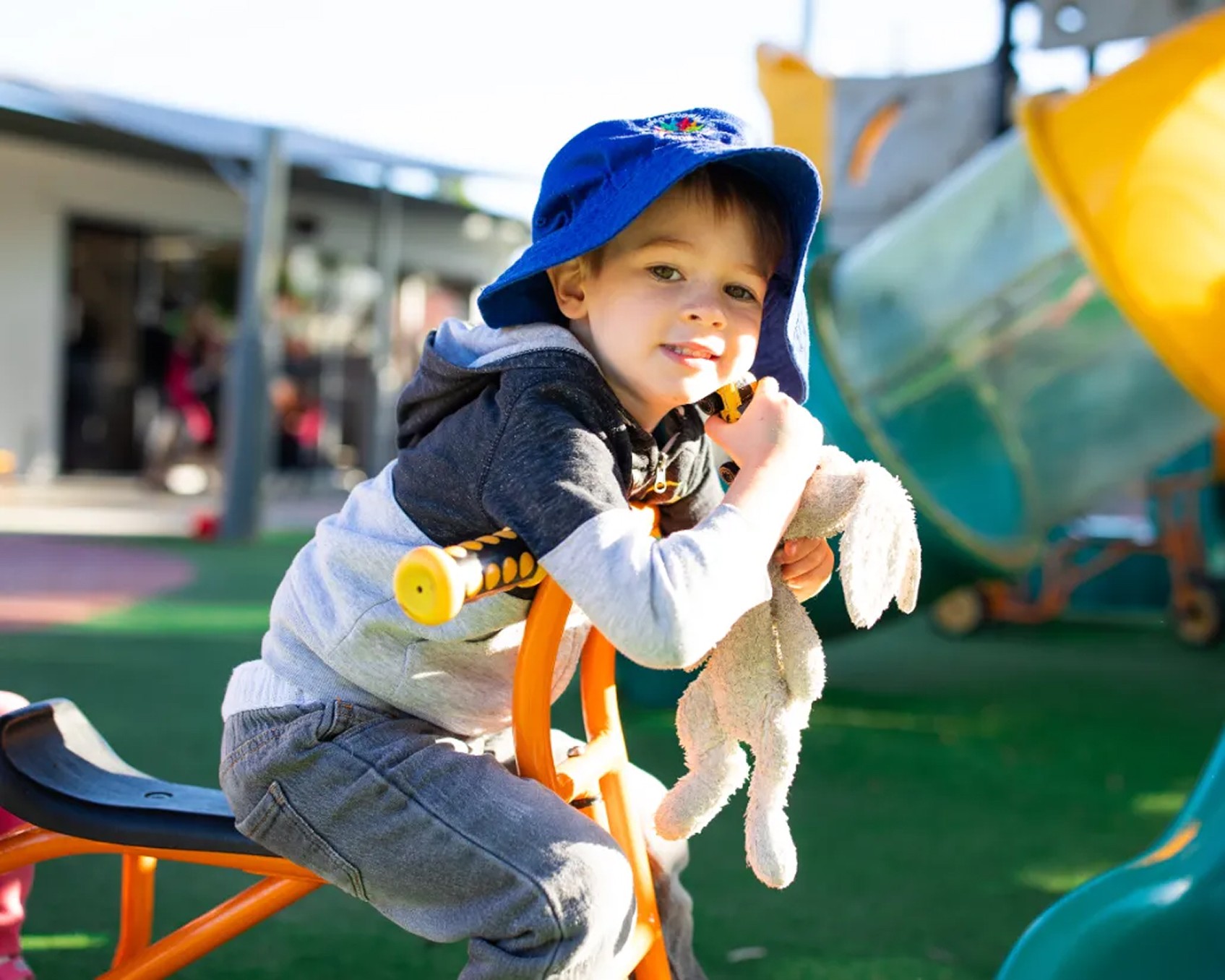 Young child playing on an outdoor tricycle while holding a soft toy at Kaleidoscope Kids childcare centre in Brisbane.