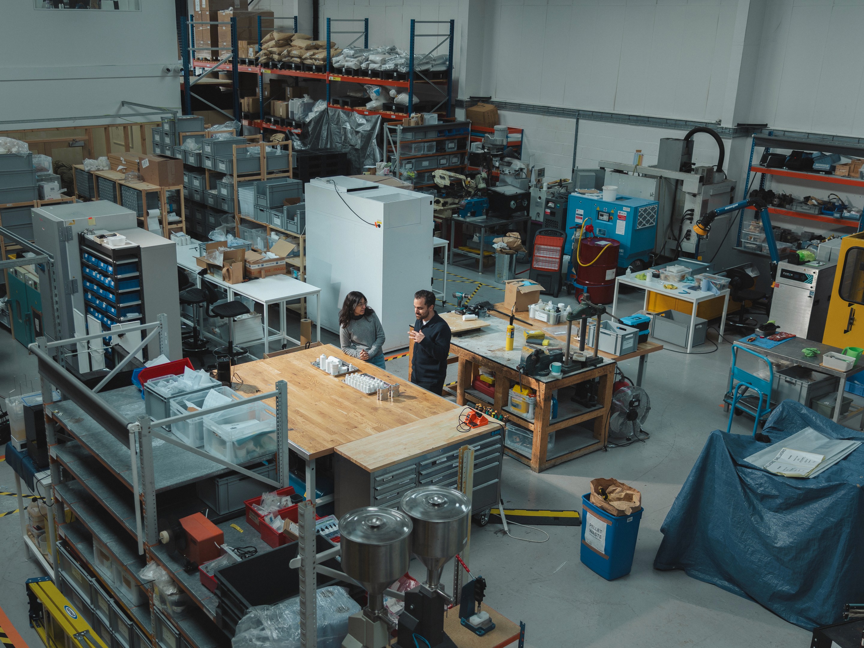 An elevated wide-angle photograph of the Shellworks workshop and R&D facility. In the centre, co-founders Insiya and Amir are discussing prototypes surrounded by injection moulders, extrusion blow moulders, workbenches, CNC machines and 3D printers. The image highlights how Shellworks manages the entire product lifecycle from initial material science formulation to scalable plastic-free manufacturing all under one roof.