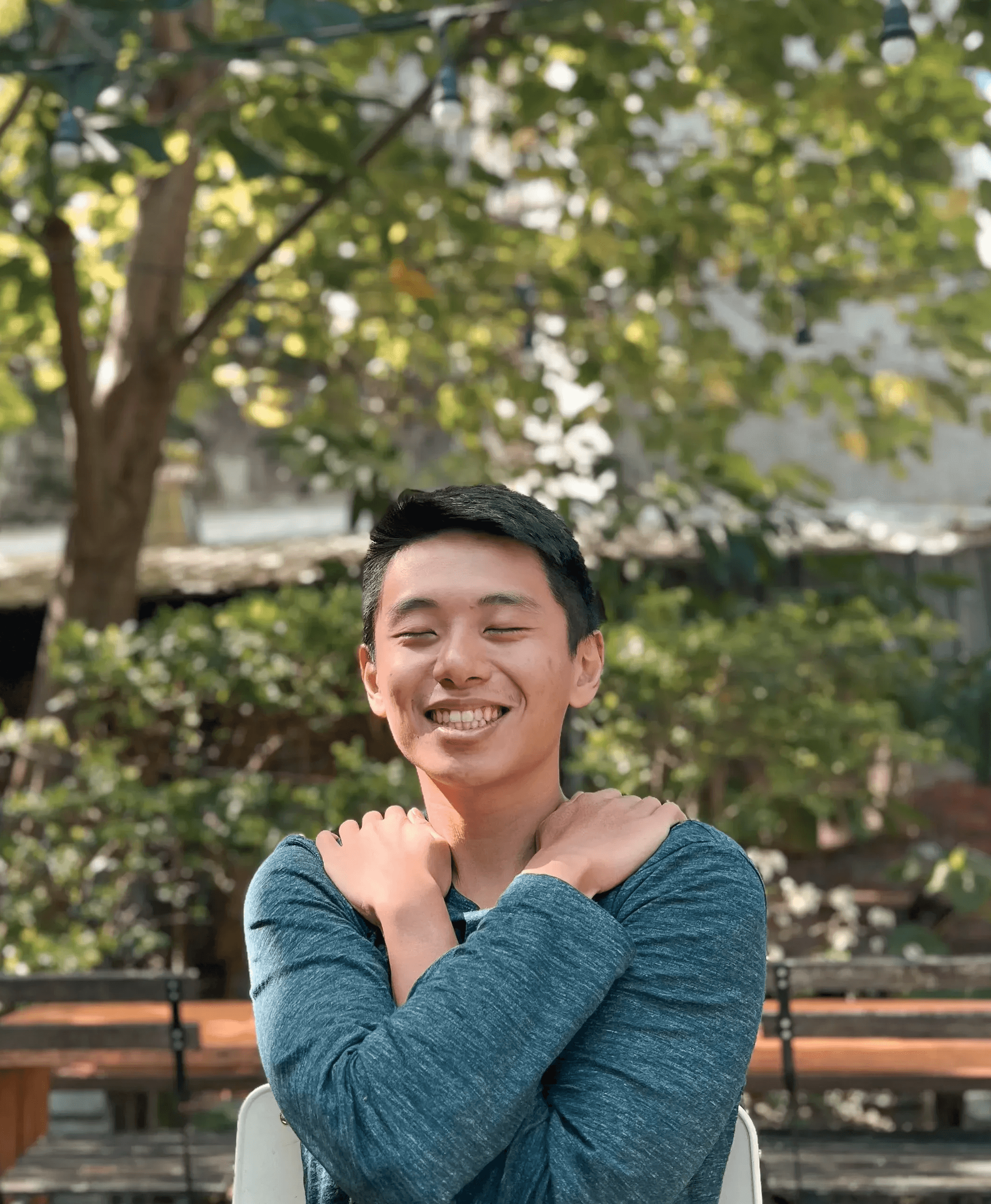 身為身心療癒引導師與「此時此地x身心沐浴」創辦人的 Sunny Chen,在台北充滿綠意的戶外空間中沐浴陽光,閉目微笑並擁抱自己,展現全然的身心放鬆狀態。