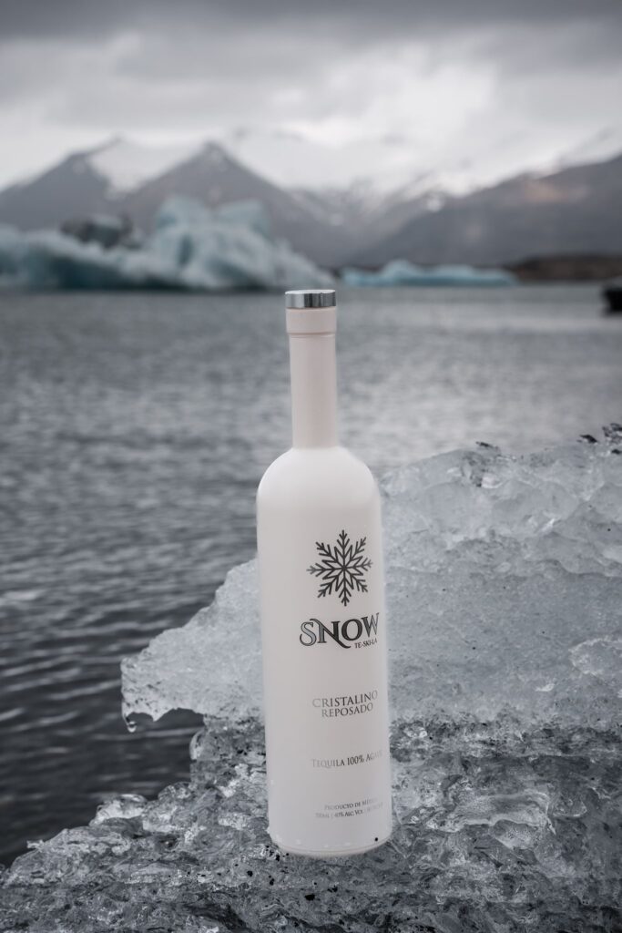 White Snow Tequila bottle resting on ice beside dark water and snow-covered mountains