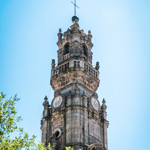 A historic stone clock tower with ornate details under a clear blue sky, with tree branches visible in the foreground.