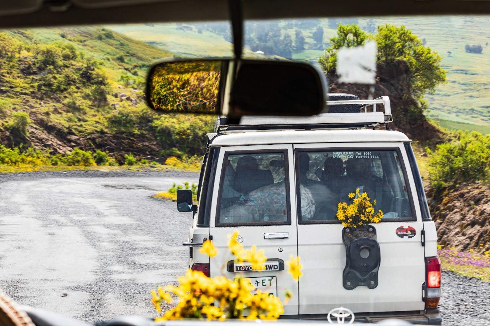 A picture of the back of a white 4x4 van full of people, decorated with yellow adey abeba flowers; the photo is taken from the driver's seat of the car behind.