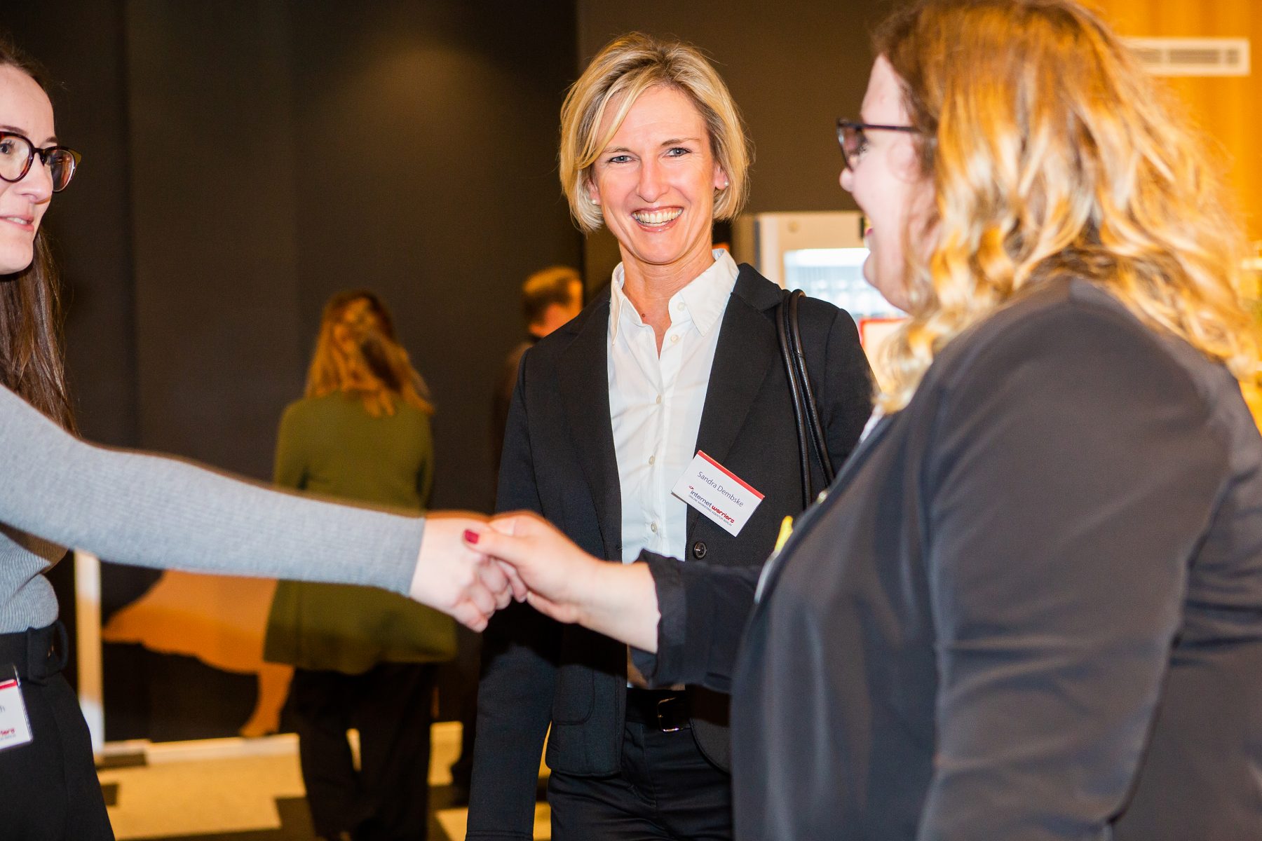 Foto: leererZwei Mitarbeiterinnen beim Kennenlernen und eine dritte Mitarbeiterin im Hintergrund - internetwarriors GmbH Konferenzraum mit Stehtischen und Großplakaten - internetwarriors GmbH