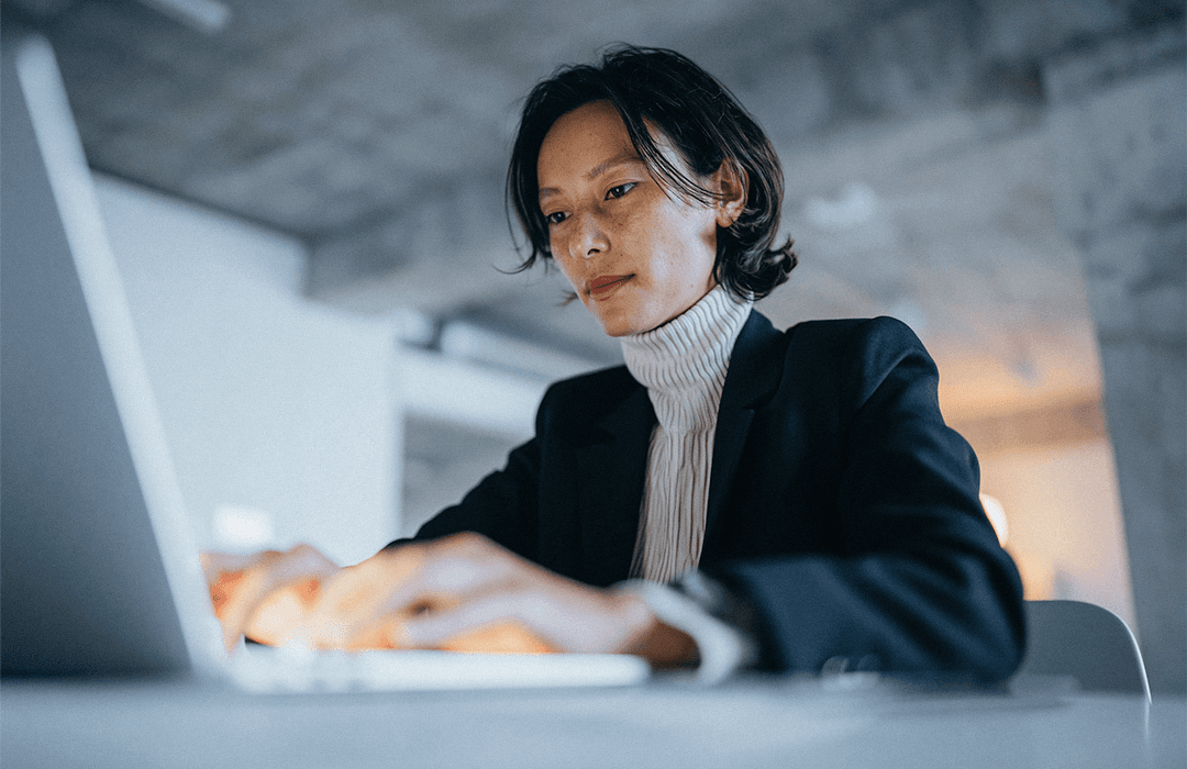 Uma mulher focada, de gola alta branca e blazer preto, digitando em um laptop em um escritório com iluminação quente. Blog post Inspira.