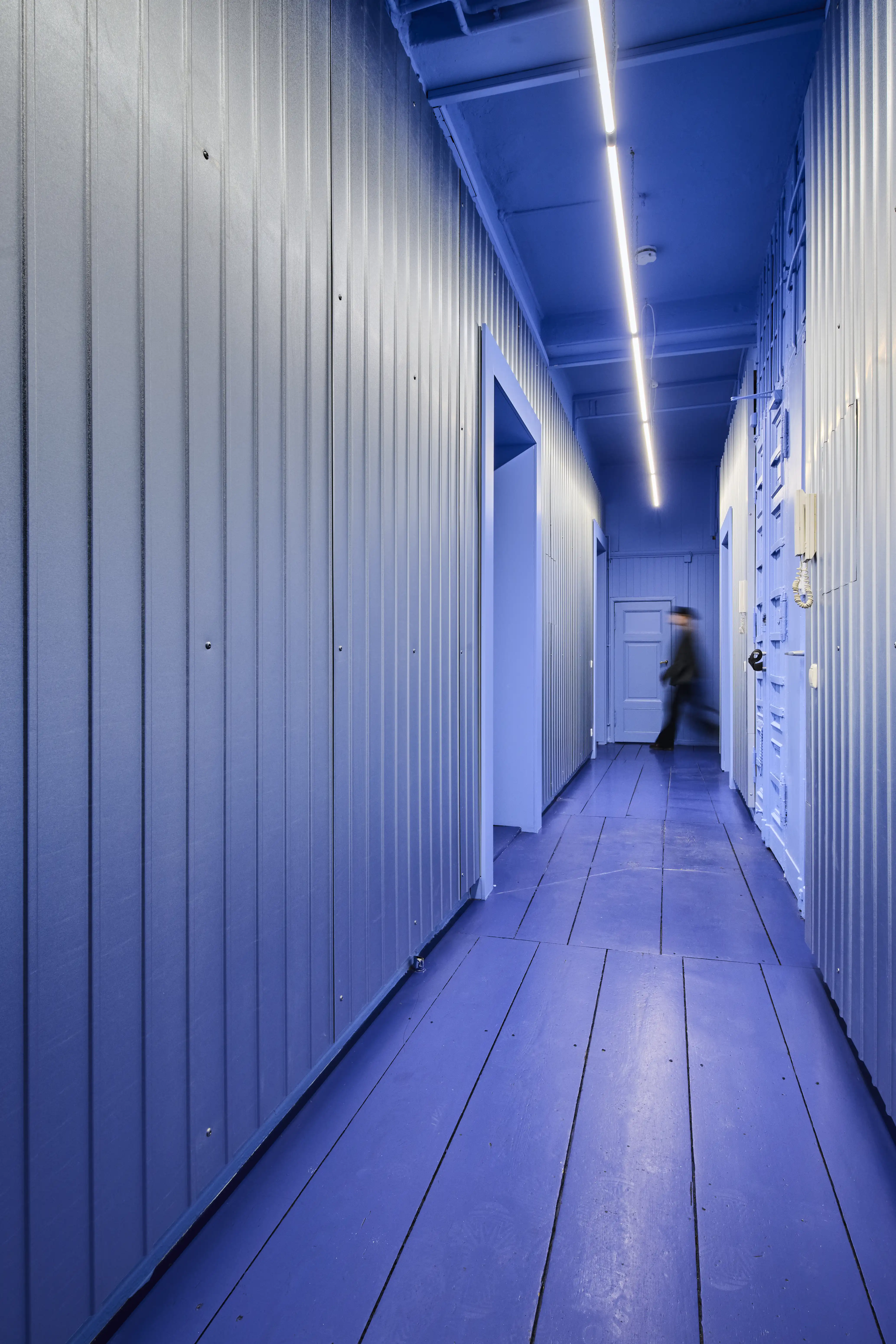 Blue-violet corridor with vertical wall panelling, plank floor and a person walking at the far end.