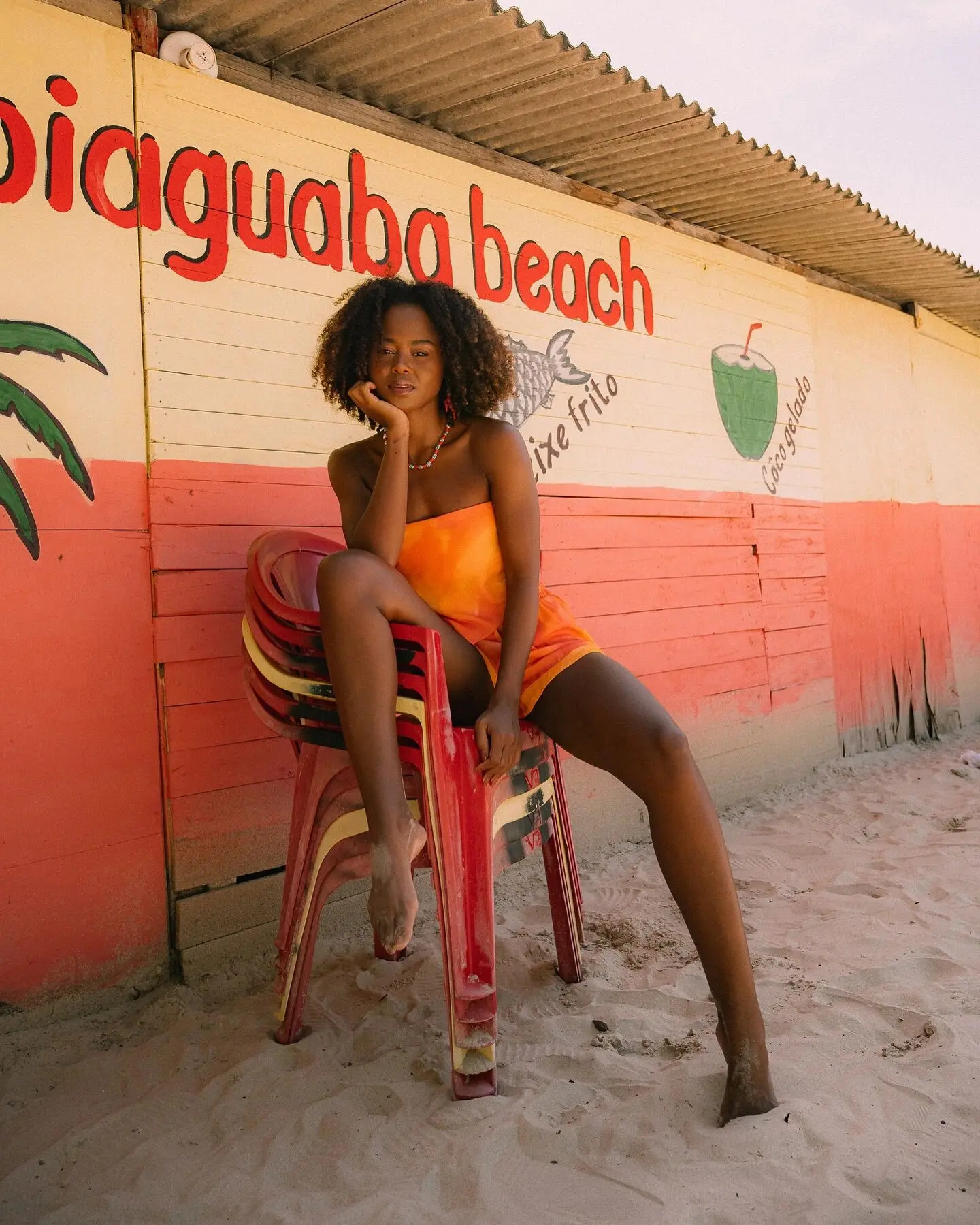 Big Jerk’s girlfriend sitting on stacked red chairs outside a painted beach shack