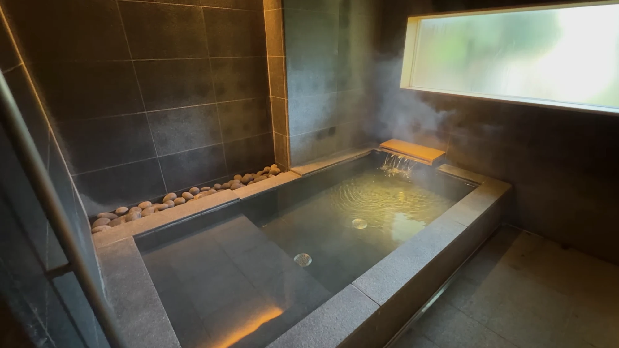 A dark stone bath in a contemporary Kinosaki inn lit by a long window above the water.