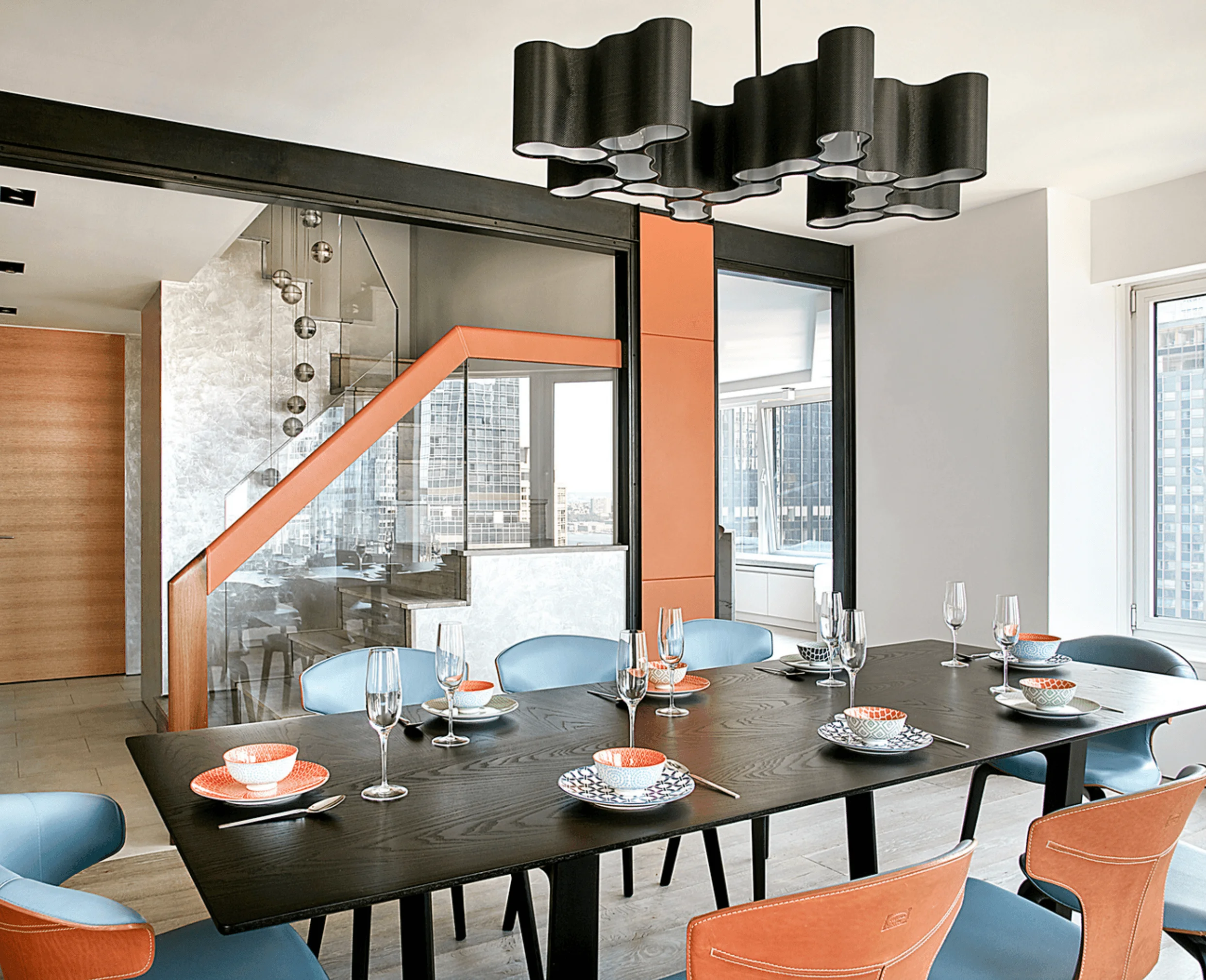 A dining table set with patterned tableware beneath a wavy black pendant light, with a glass staircase and city views beyond