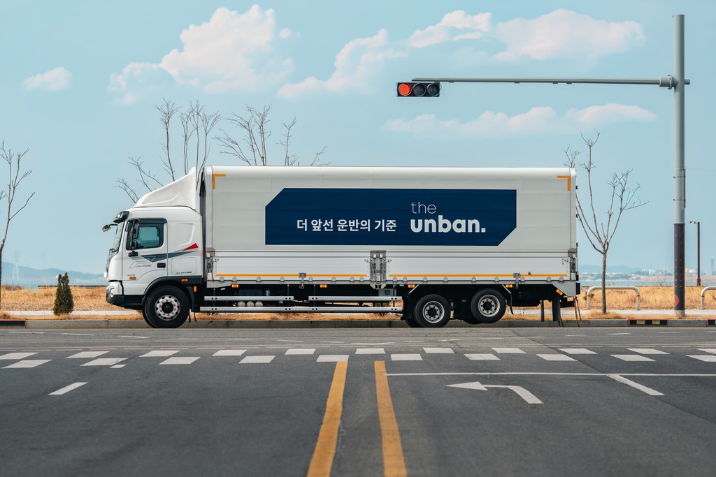 CJ대한통운 더 운반 브랜드 로고가 래핑된 대형 화물 운송 차량이 도로 위에 정차해 있는 모습입니다. '더 많은 운반의 기준'이라는 문구와 함께 더 운반의 운송 서비스 정체성을 시각적으로 표현하고 있습니다.