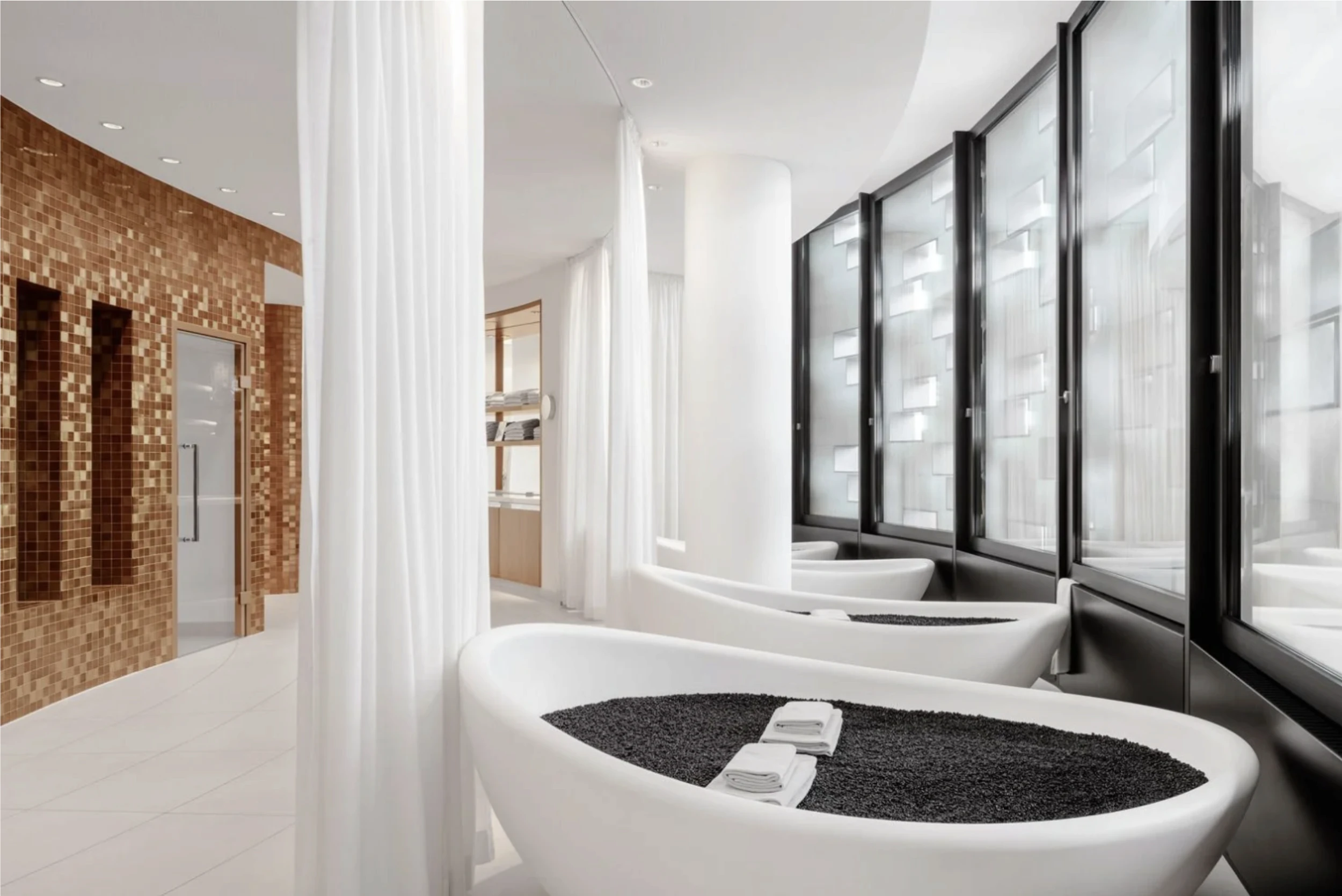 Modern spa with soaking tubs filled with black stones beside large windows and white curtains