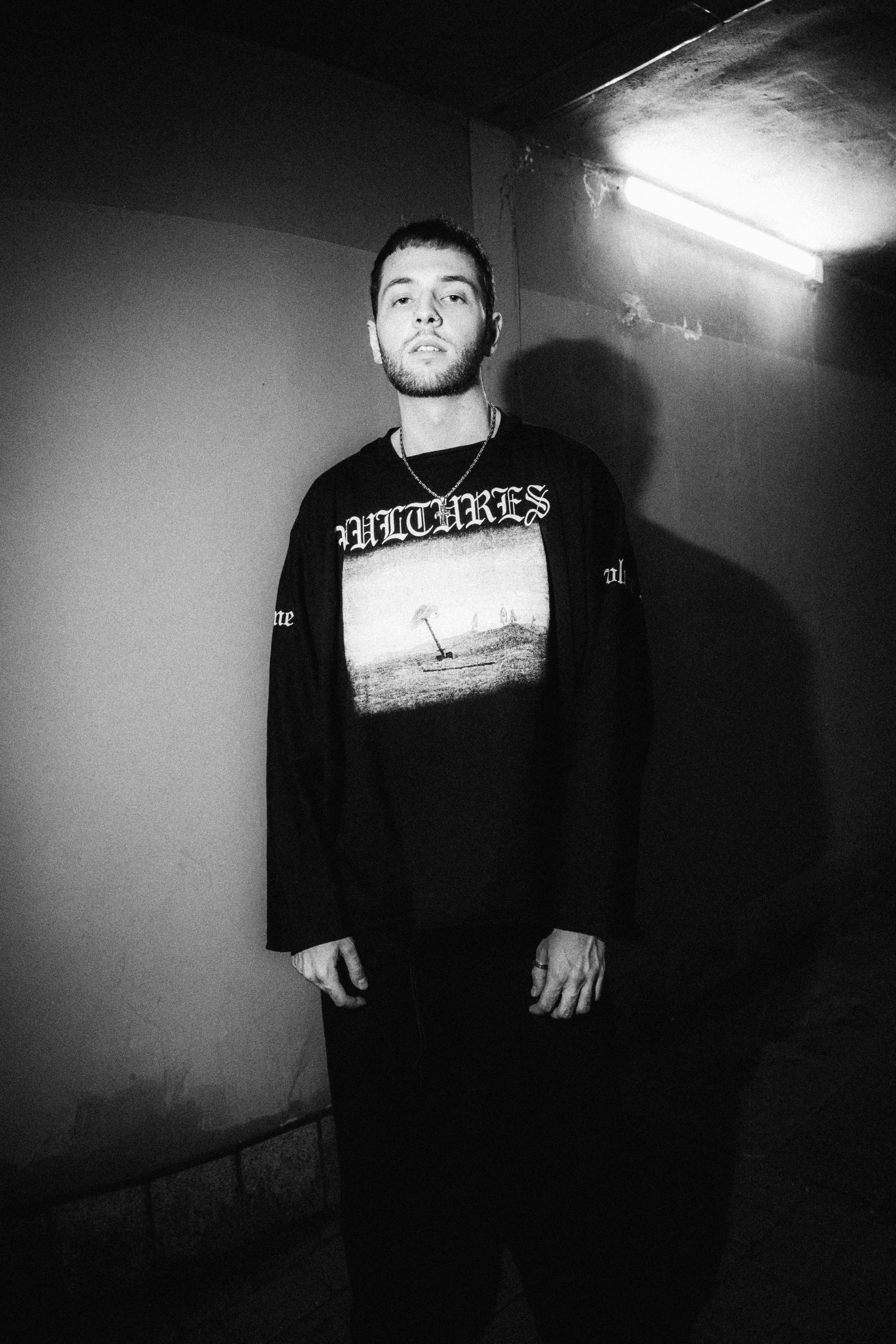 Black and white portrait of SHDØW, a Polish music producer, standing against a dimly lit wall. SHDØW is wearing a long-sleeve shirt featuring artwork and the word 'VULTURES,' referencing his work on Kanye West and Ty Dolla $ign's Vultures 1 and 2.