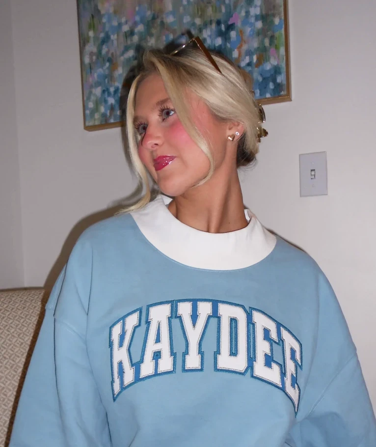 Indoor photo of a person wearing a light blue “KAYDEE” crewneck with a white mock-neck collar, looking off to the side.