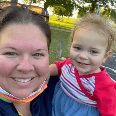 Portrait of Julie Wilson smiling outdoors while holding a young child, with a sunny park setting in the background.
