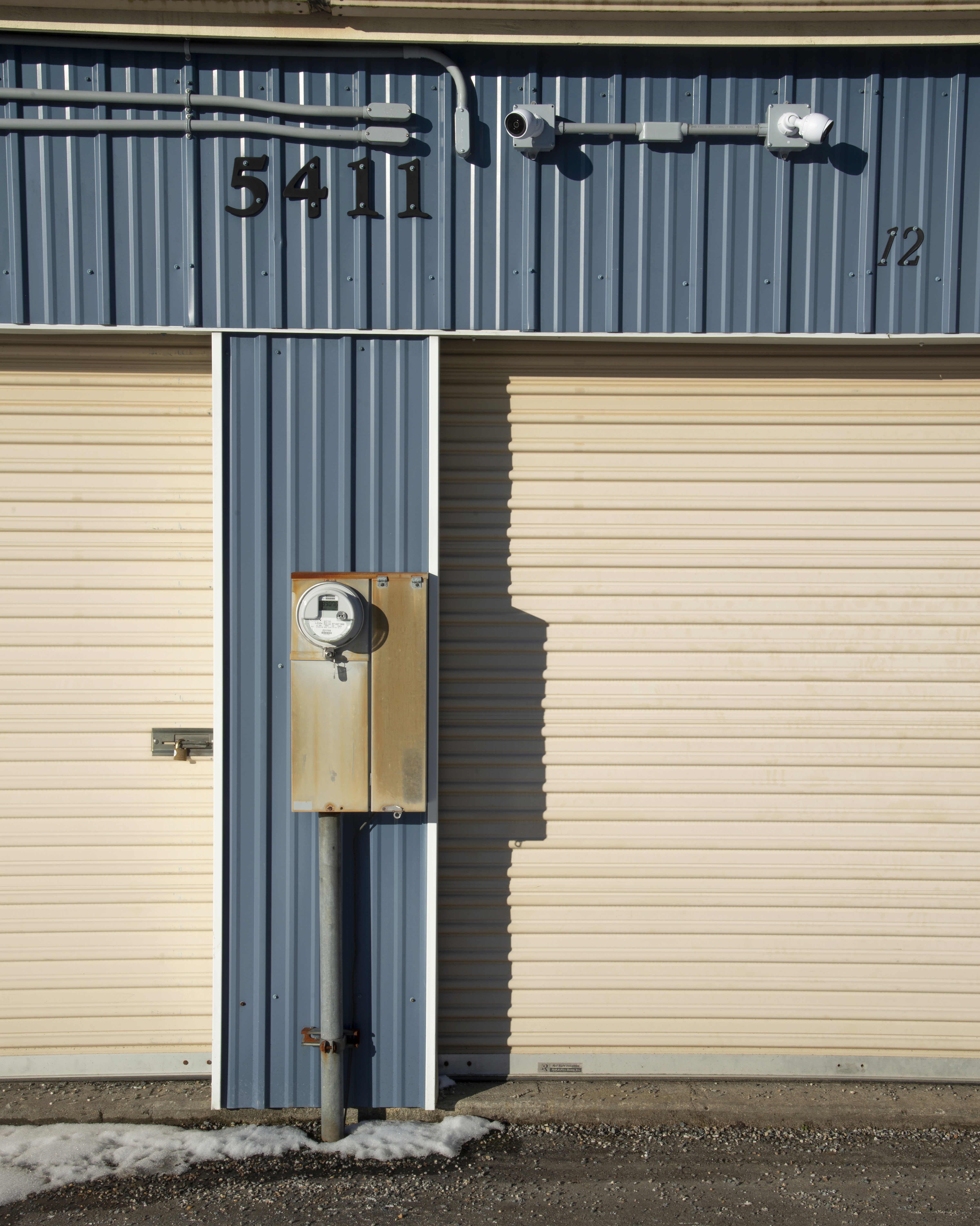 Close-up of two beige roll-up storage unit doors on a blue-grey corrugated metal building, with the unit number "5411" stenciled above and a utility box mounted between the doors on a concrete pad with light snow at the base.