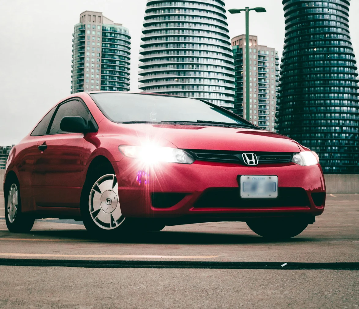 Une Honda Civic rouge photographiée sur un toit-terrasse, face à des immeubles torsadés contemporains. Le contraste entre le design simple de la compacte et l’architecture futuriste crée une scène dynamique et urbaine.