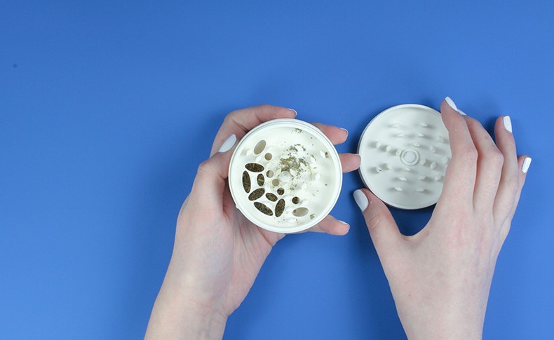 Two hands with white-painted nails hold a white ceramic bowl containing white yogurt topped with dark seeds and a sprinkle of green herbs. Beside it lies a white ceramic strainer with small holes. Both items rest on a blue surface.