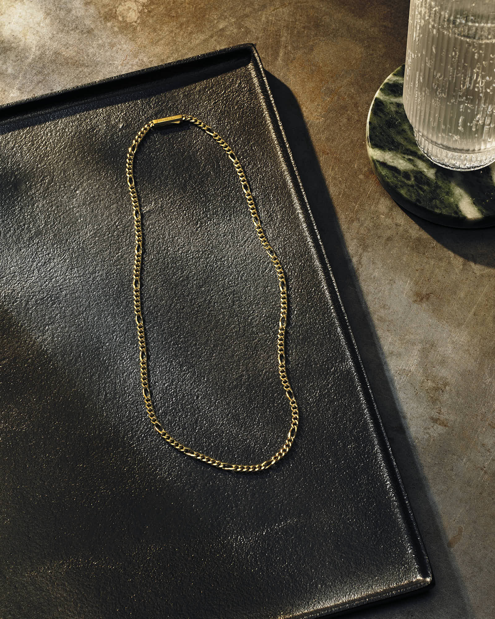 Gold chain necklace on black tray with glass of water nearby.