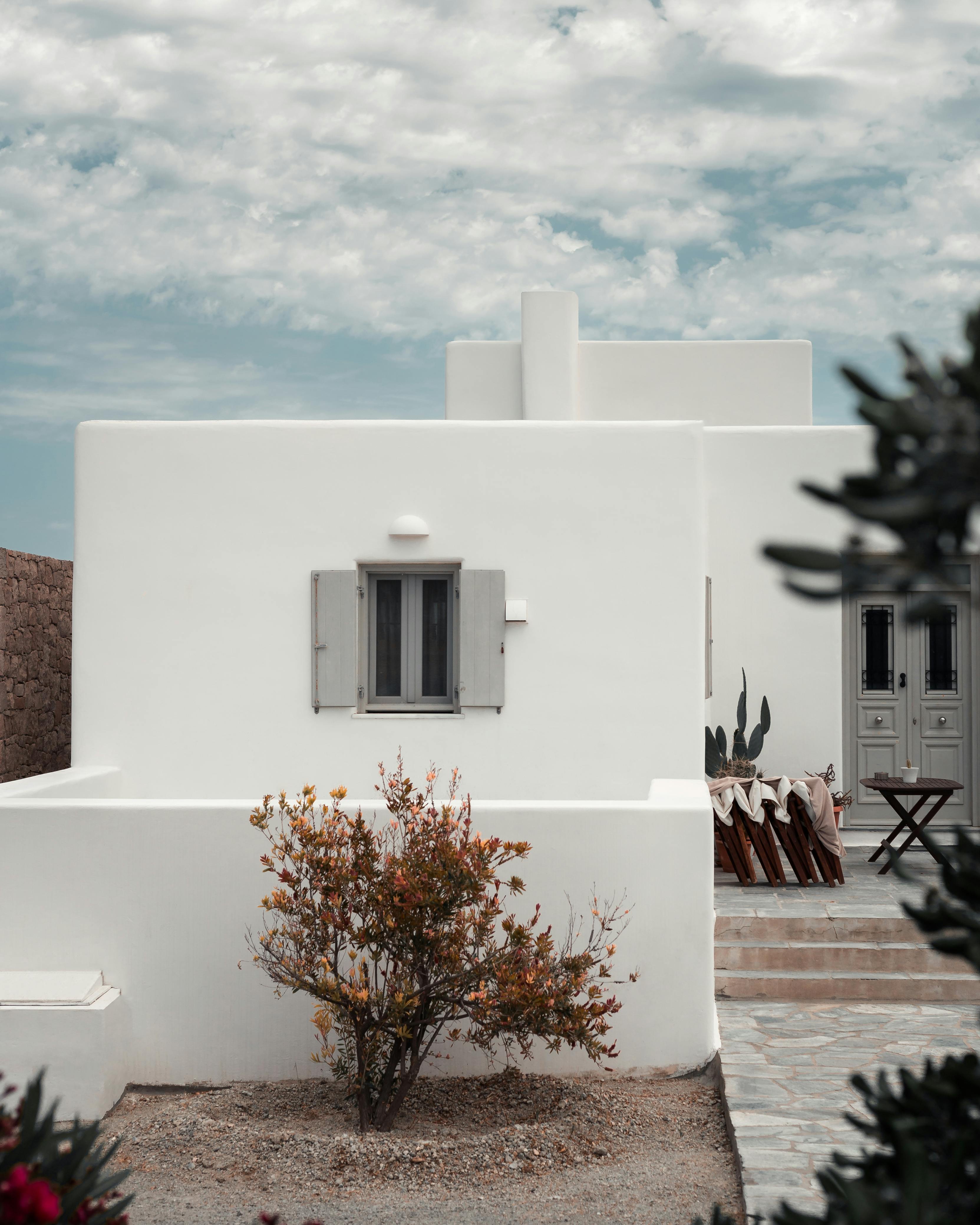 A picturesque whitewashed building with minimalist architecture stands under a partly cloudy sky, framed by a lush plant and rustic outdoor furniture, capturing the serene ambiance of a Mediterranean landscape.