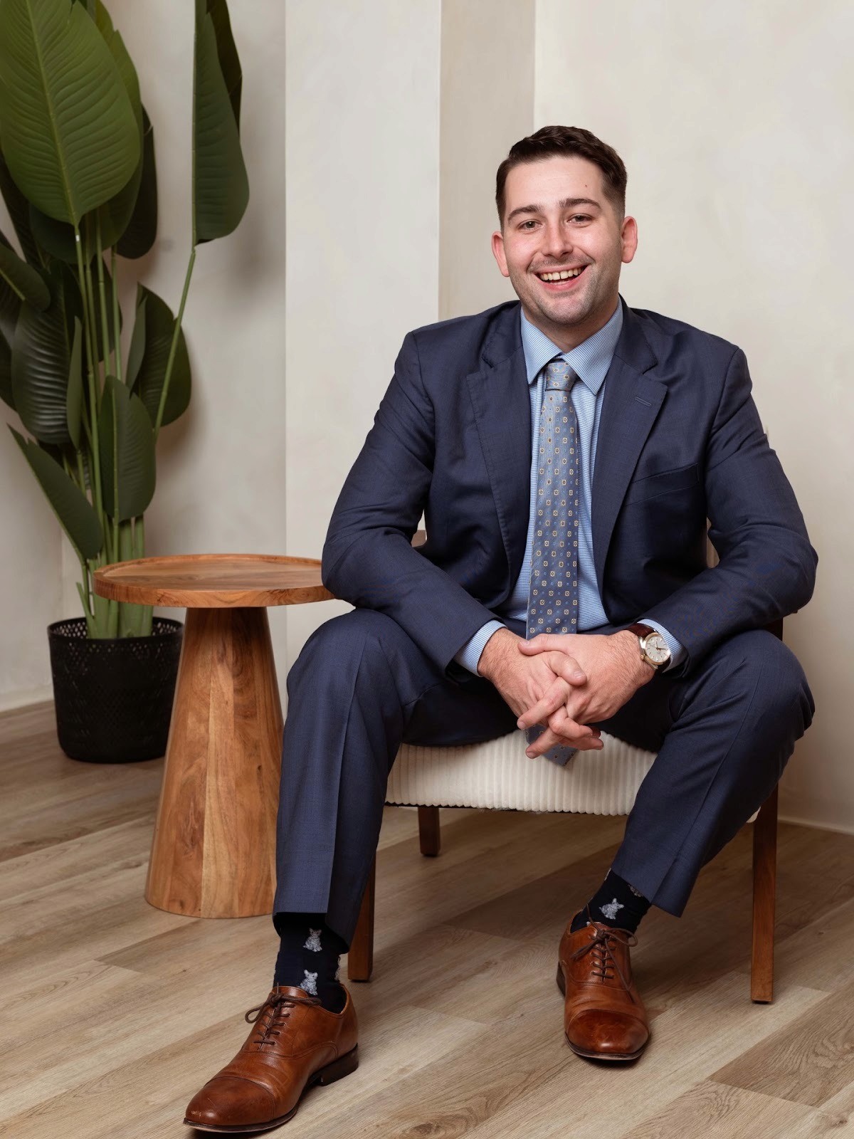 Man in a blue suit and tie, smiling while seated on a chair. He wears brown shoes and star-patterned socks, with a plant and wooden table nearby.