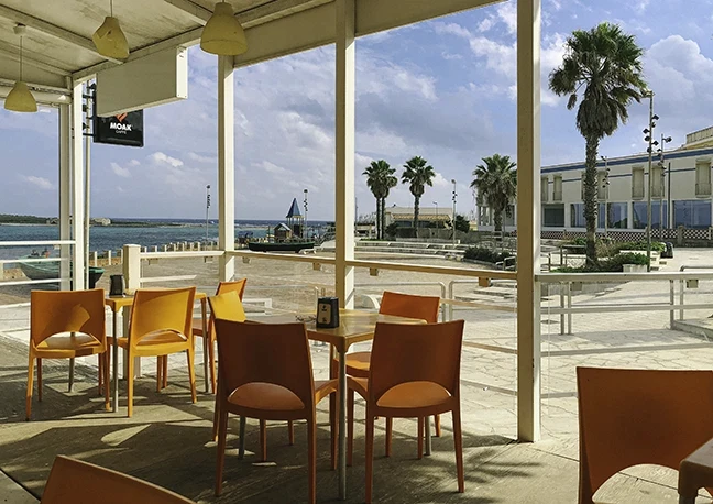 Letnia kawiarnia z pomarańczowymi krzesłami, widok na morze i palmy w tle, jasne światło i nadmorska atmosfera