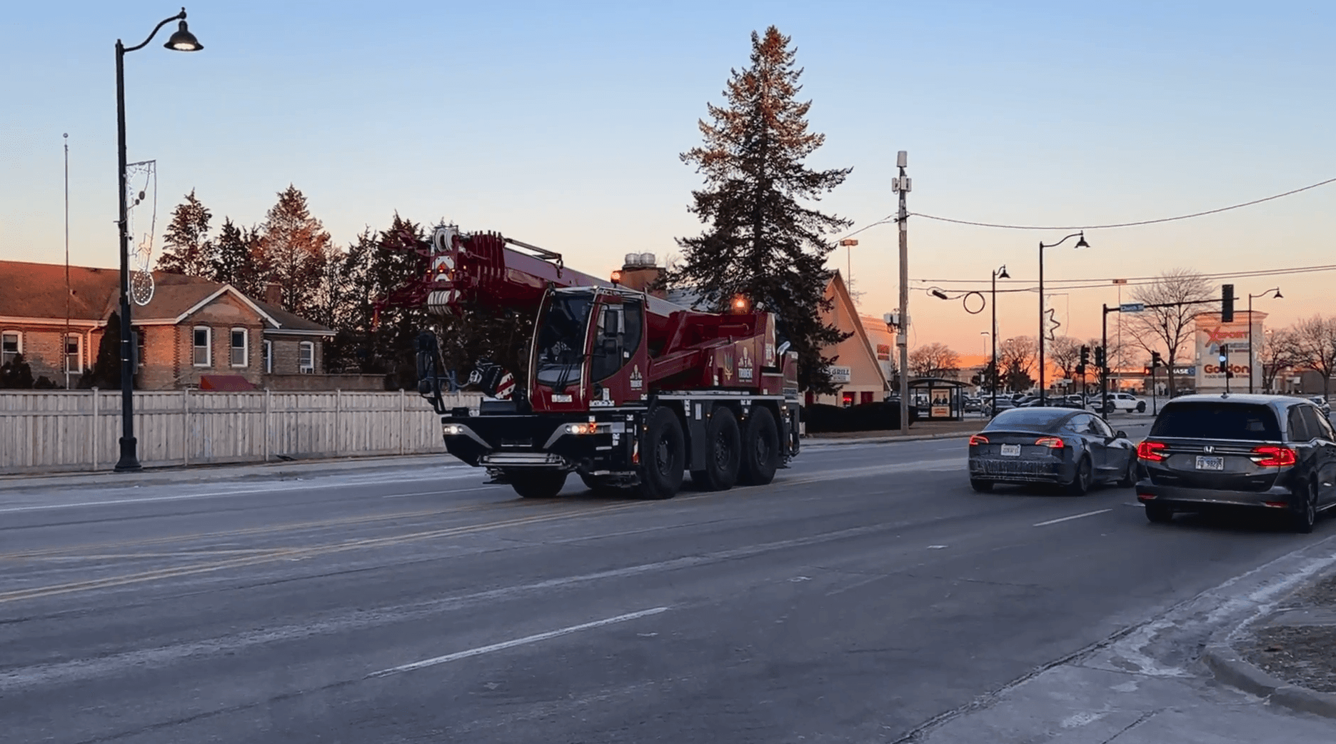 A red crane truck drives down a suburban street at sunset, surrounded by parked cars and lined with small trees and lamp posts.