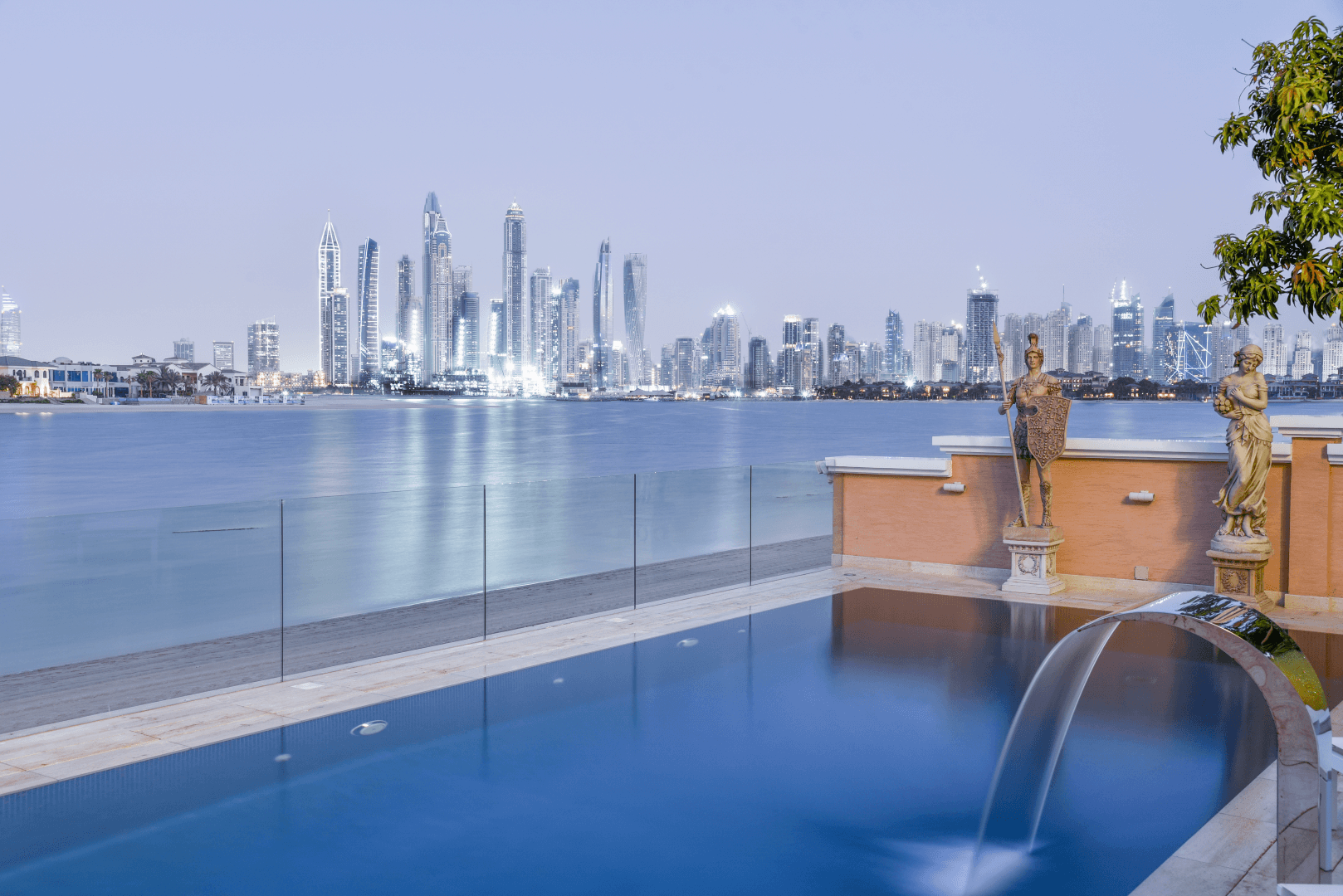 A luxurious infinity pool overlooks a serene waterfront, with a backdrop of a modern city skyline lit up against the evening sky, featuring iconic skyscrapers and elegant statues adorning the poolside.