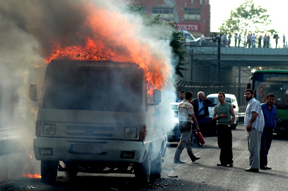 A van engulfed in flames on a city street with bystanders observing the scene.