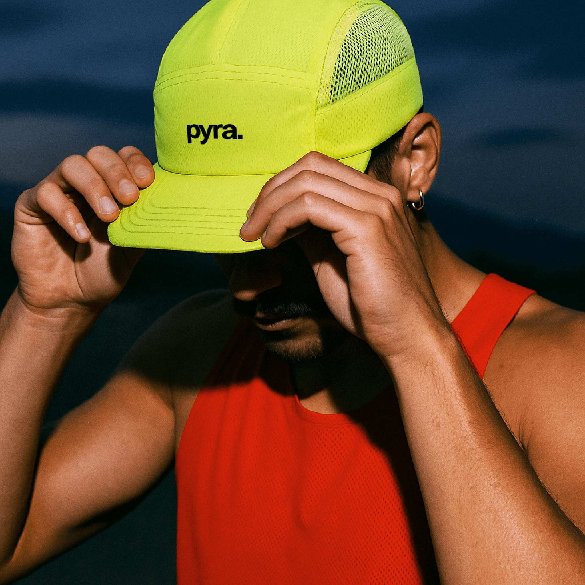 Man wearing a neon yellow cap with "pyra." logo and a red athletic tank top, adjusting his hat with both hands.