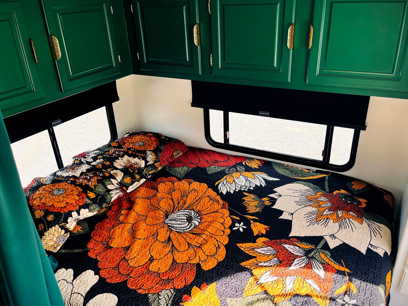 RV interior bedroom with large floral quilt featuring orange and white flowers on a dark background. Overhead are green cabinets with gold handles.