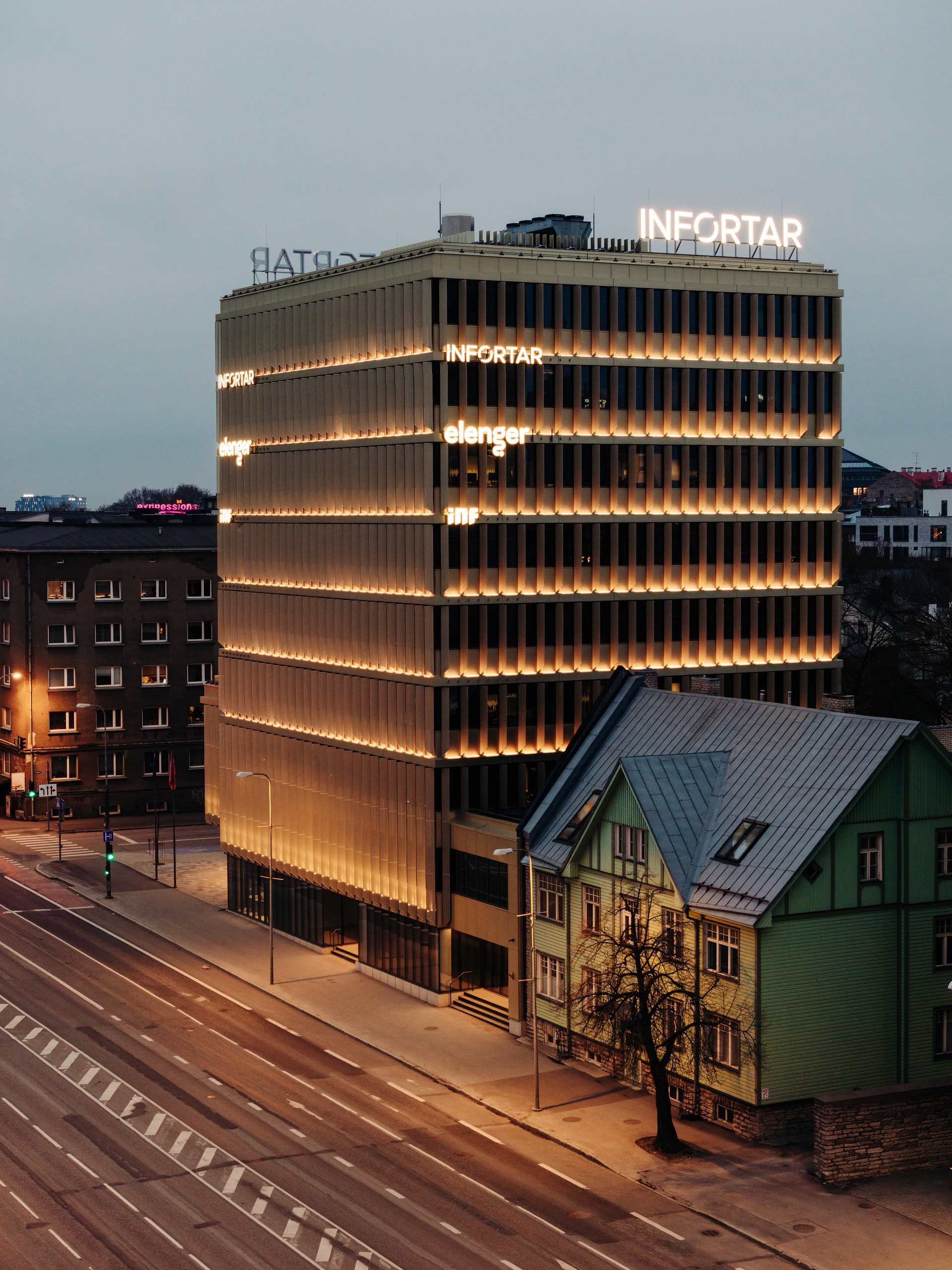 Vertical evening view of the full illuminated building on a street corner, with the adjacent green gabled house and quiet road in the foreground.