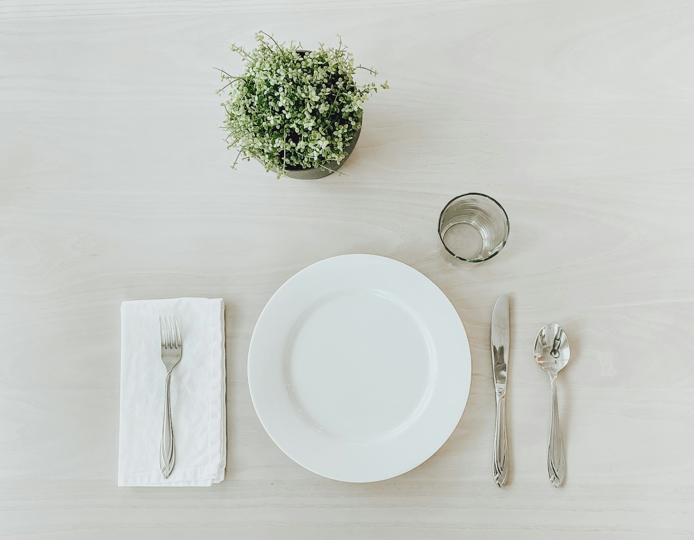 A simply set place with a small white plate, folded napkin, water glass, and cutlery on a light table, illustrating smaller, slower meals that ease slowed digestion.