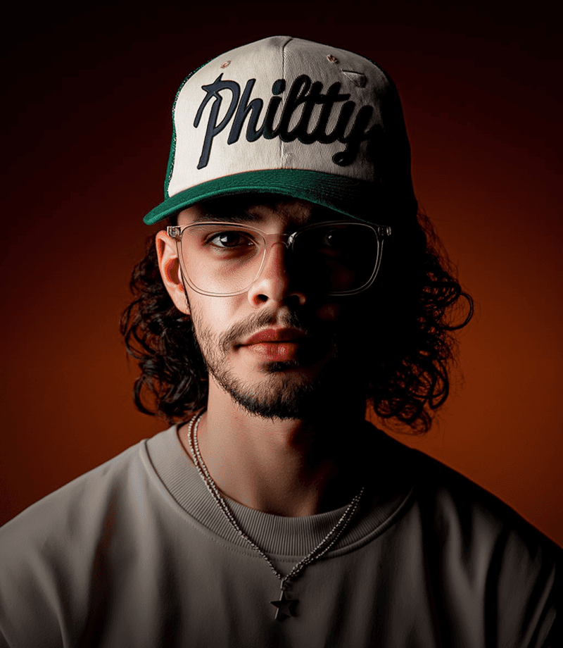 A young man with round glasses, a nose ring, and facial hair looks toward the camera against a dark, softly colored background.