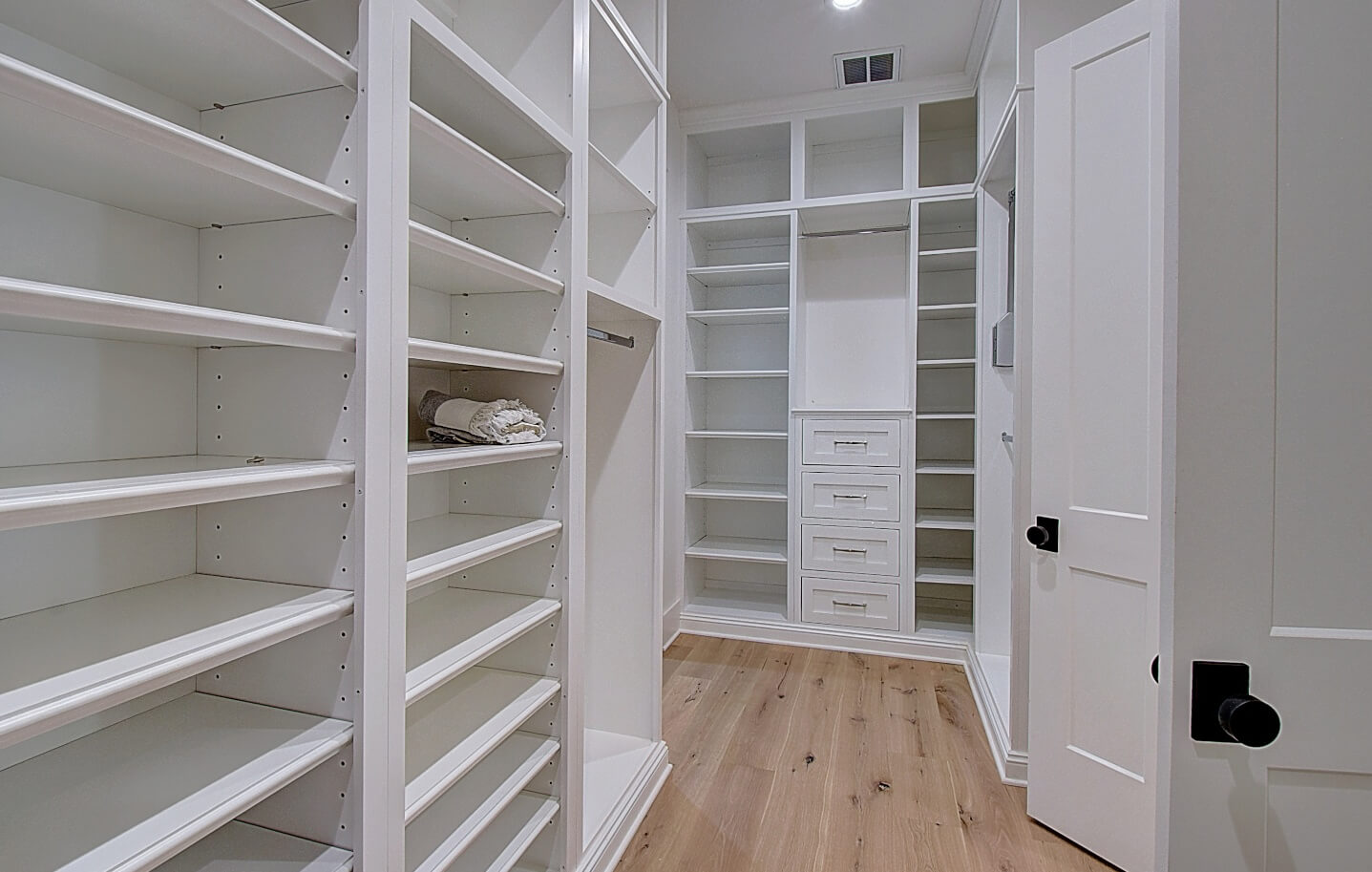 A spacious, modern bedroom walk in closet with white shelves and a door, featuring light wood flooring.