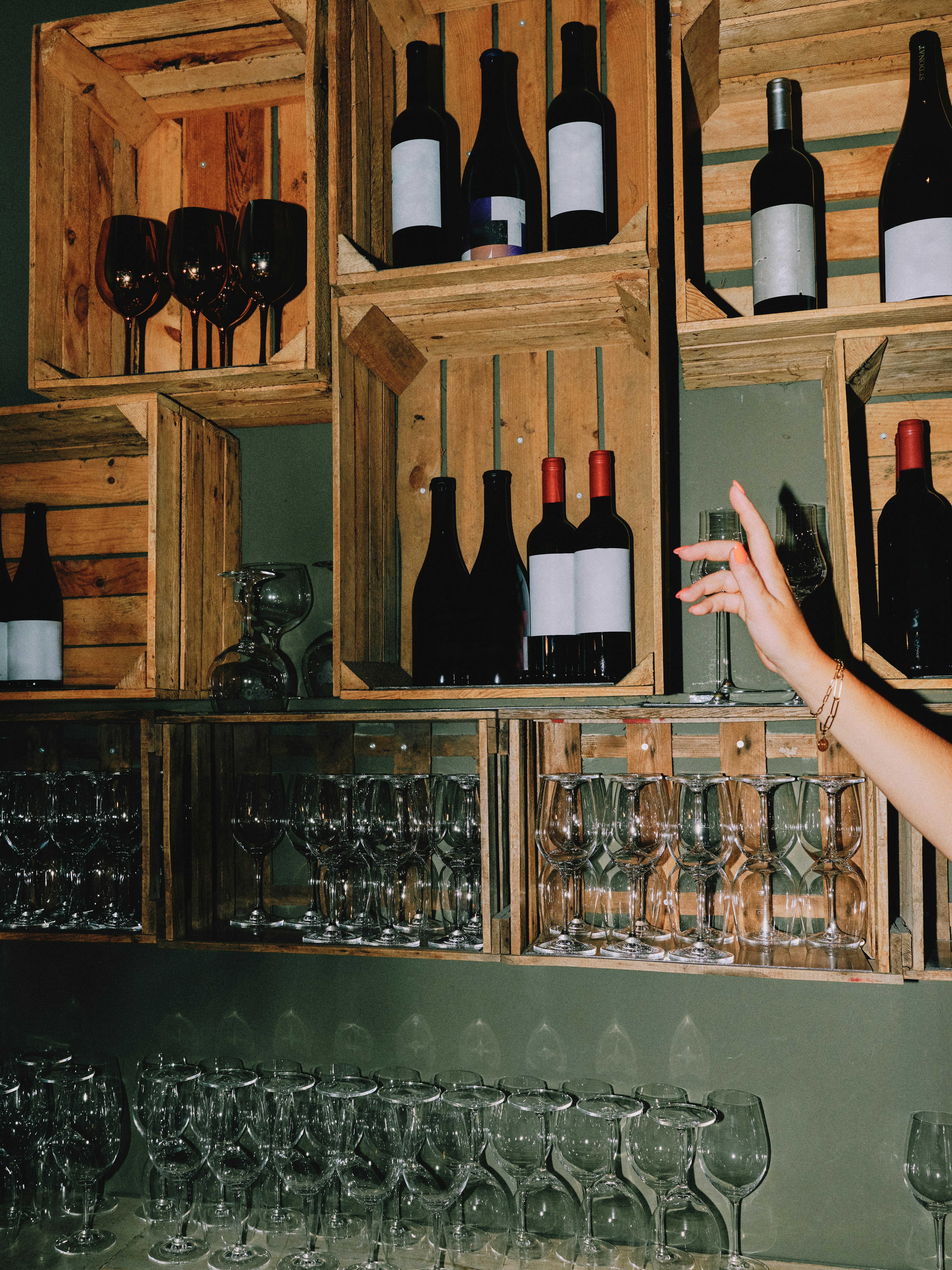 Moody interior shot of a wine bar with wooden crate shelving, rows of wine bottles and glassware, a hand reaching in from the right side