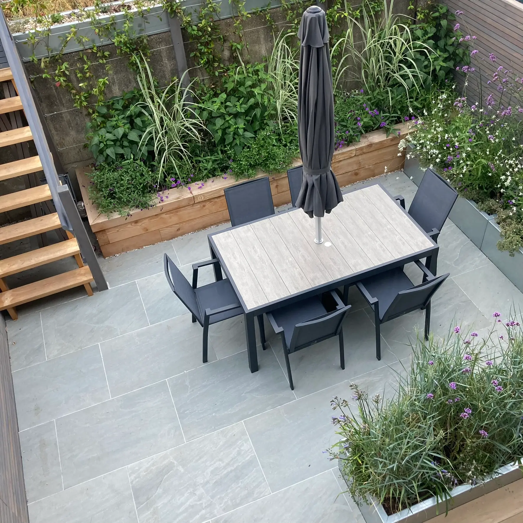 Aerial view of a modern patio with a dining table, chairs, and greenery, surrounded by wooden planters and steps.