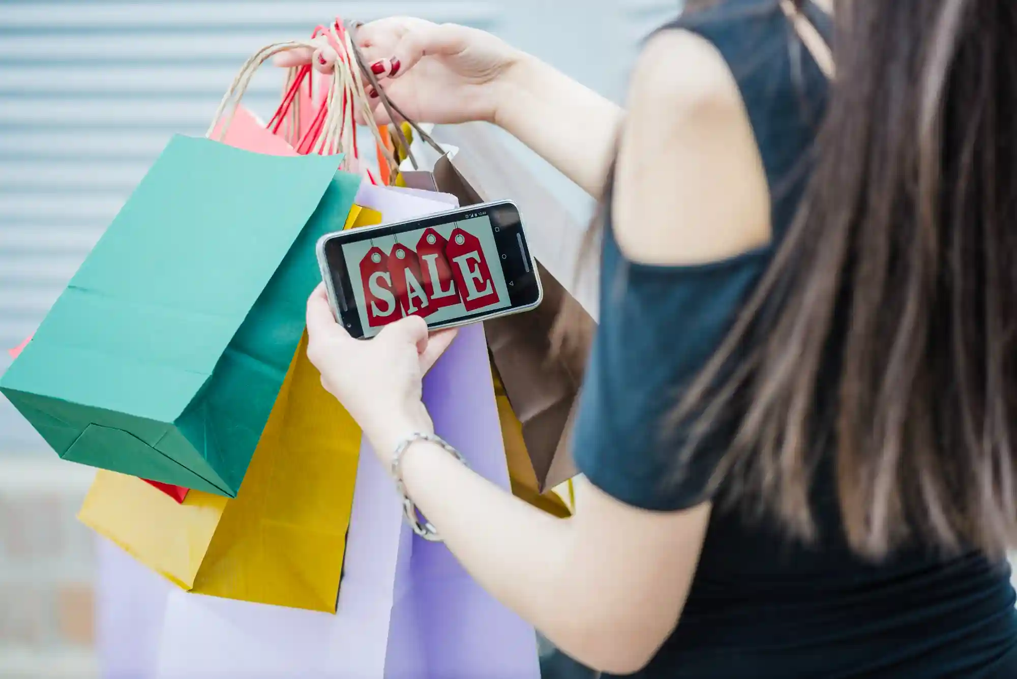 A person holds several colorful shopping bags while looking at a "SALE" notification on their smartphone screen.