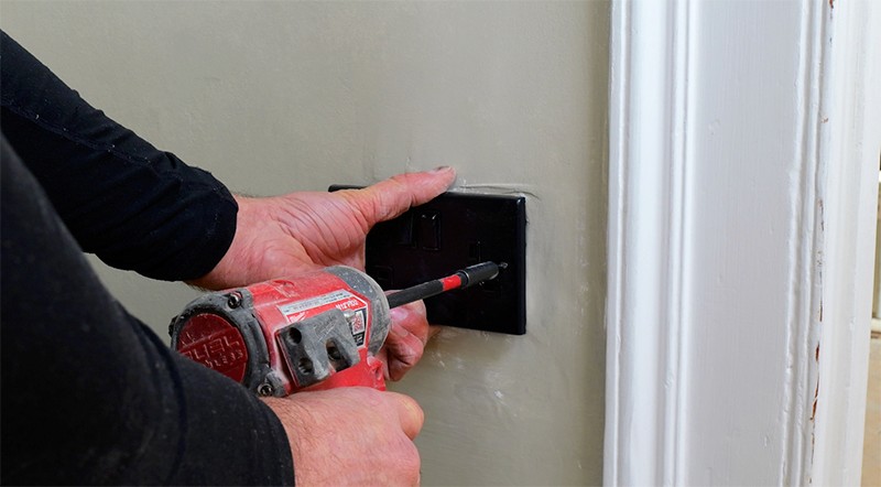 A person using a screwdriver to install or remove an electrical outlet cover on a wall.