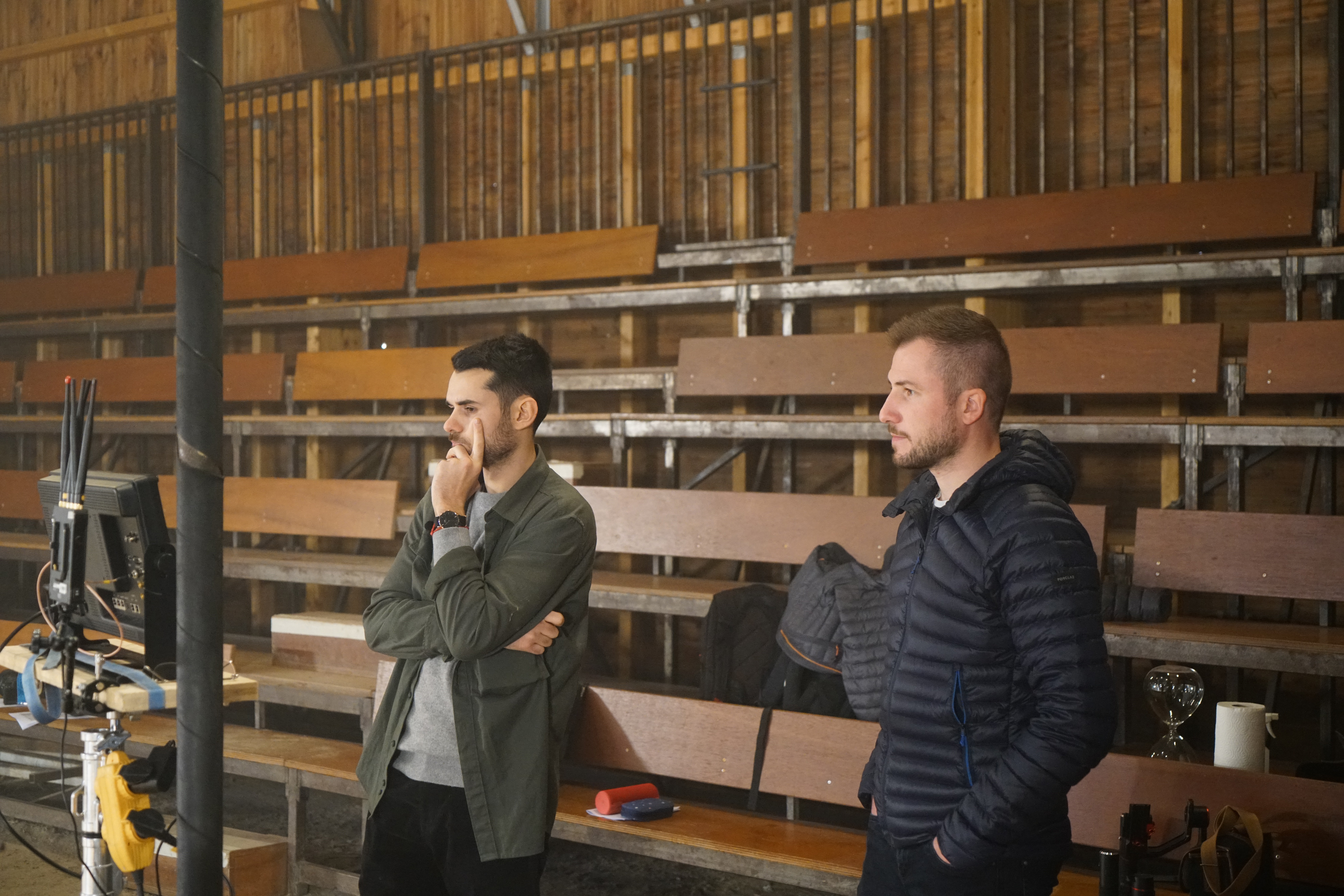 Two camera operators in a dimly lit studio adjust a large video camera. They wear headsets and caps, focusing intently. Mood is professional and focused.