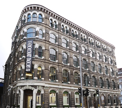 A smiling man with curly hair is sitting at a desk, looking at the camera, with a laptop and a lamp in the background.